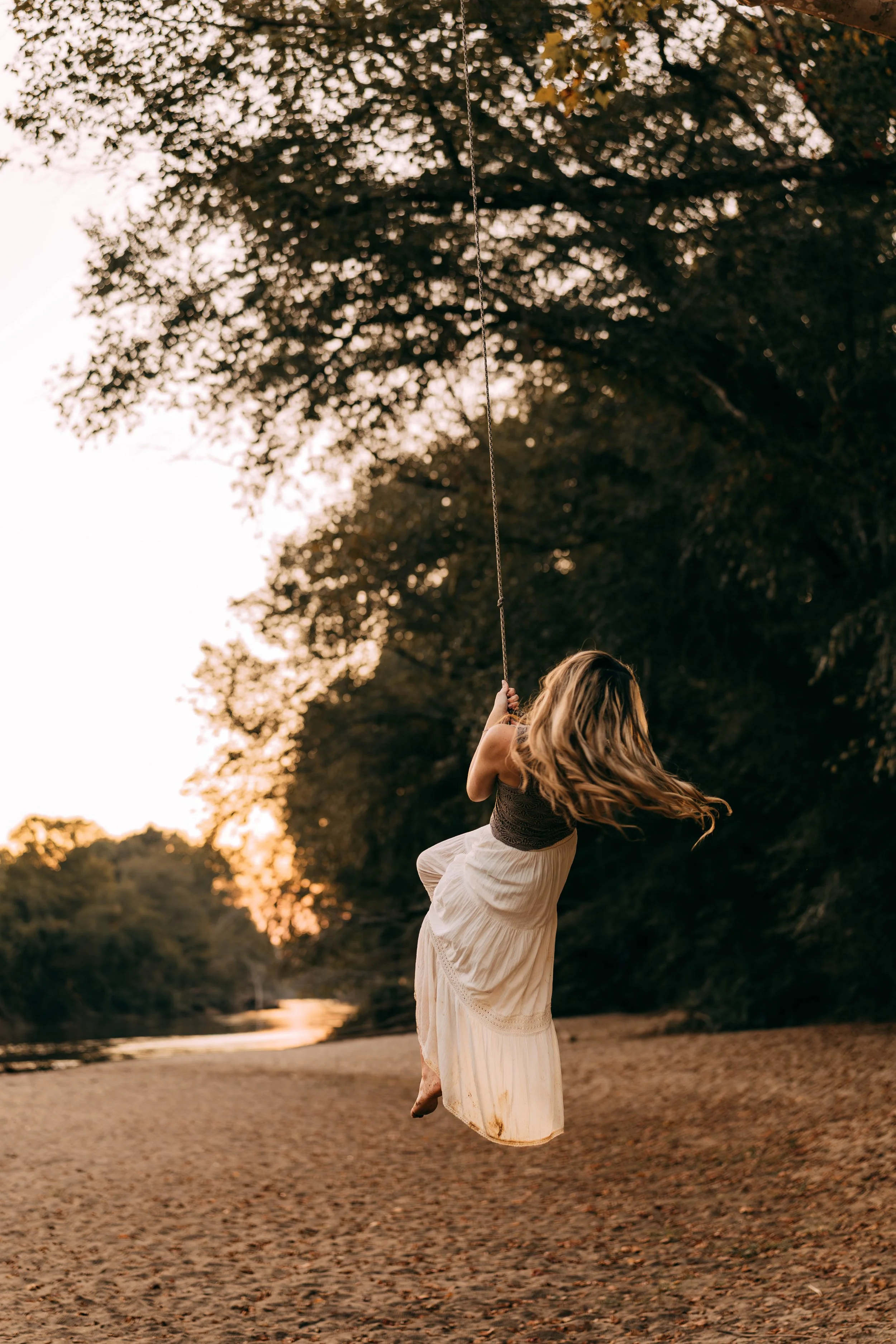 A girl wearing a dark top and light skirt swinging on a rope swing over a sandy beach at sunset, with trees and a river in the background.
