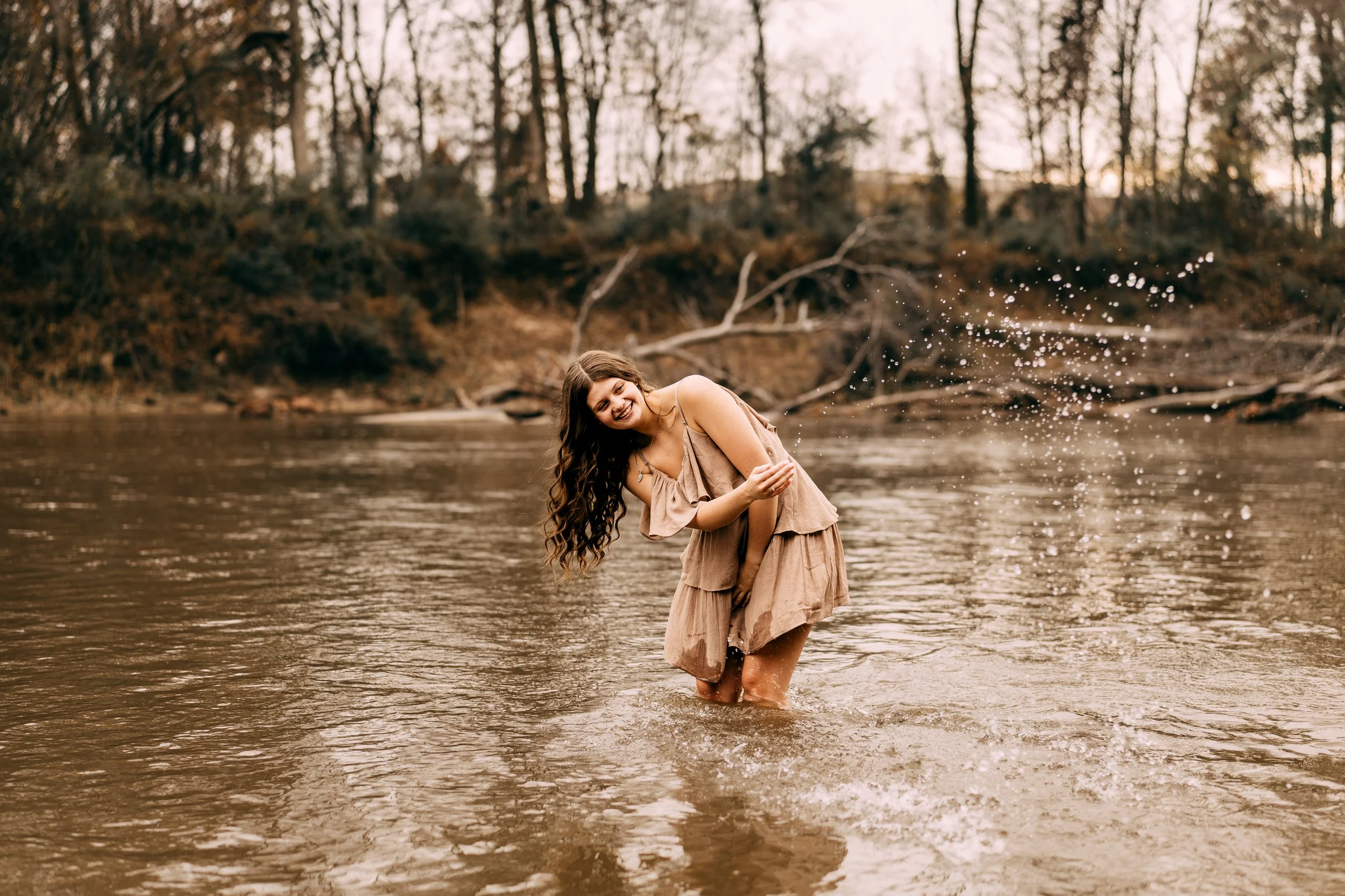 A woman wearing a beige dress standing in a river, smiling and splashing water, with trees and fallen branches in the background during sunset.