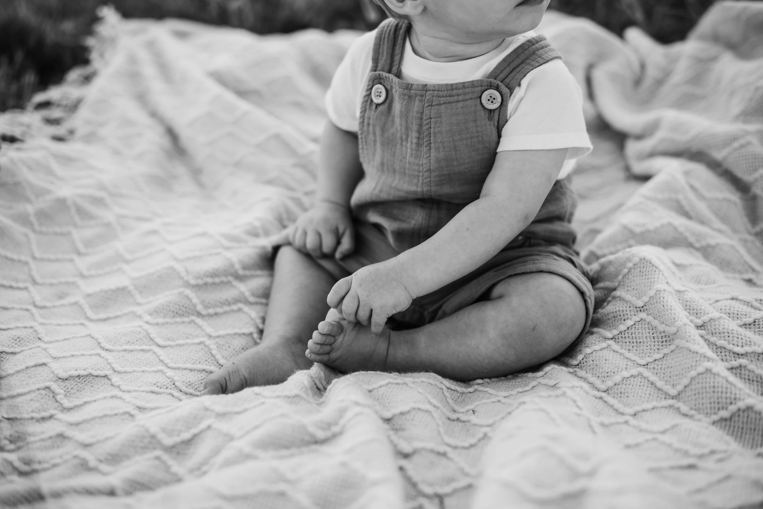 A young child sitting on a quilted blanket outdoors, with legs crossed and holding onto their foot.