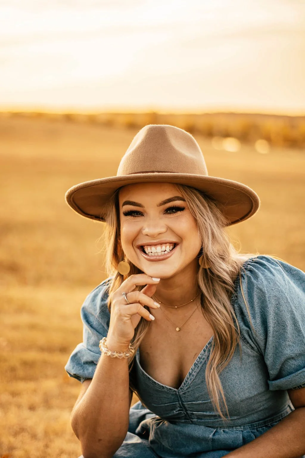 Smiling woman wearing a beige hat and denim dress outdoors during sunset.