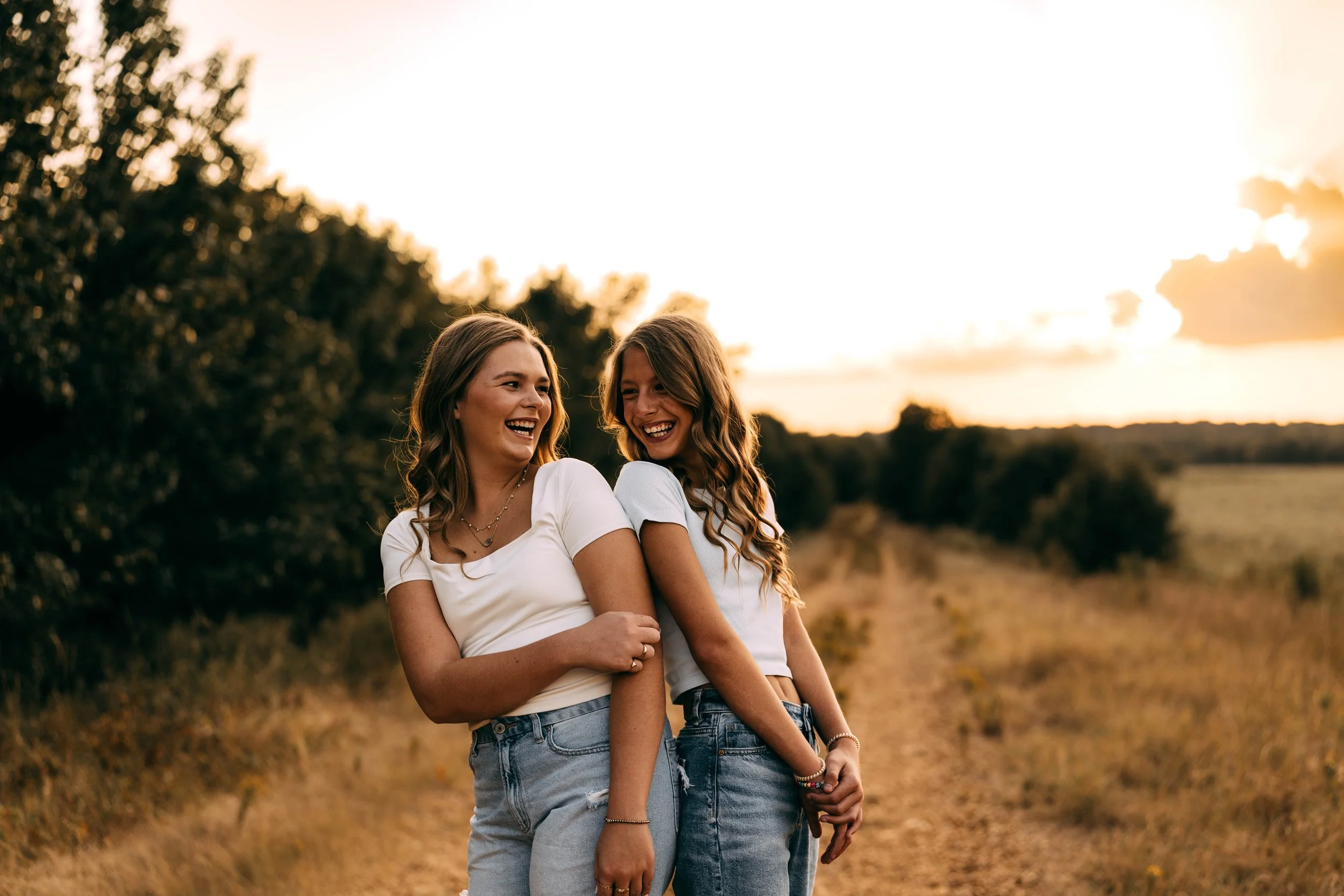Two women smiling and laughing on a dirt path during sunset, surrounded by trees and open fields.
