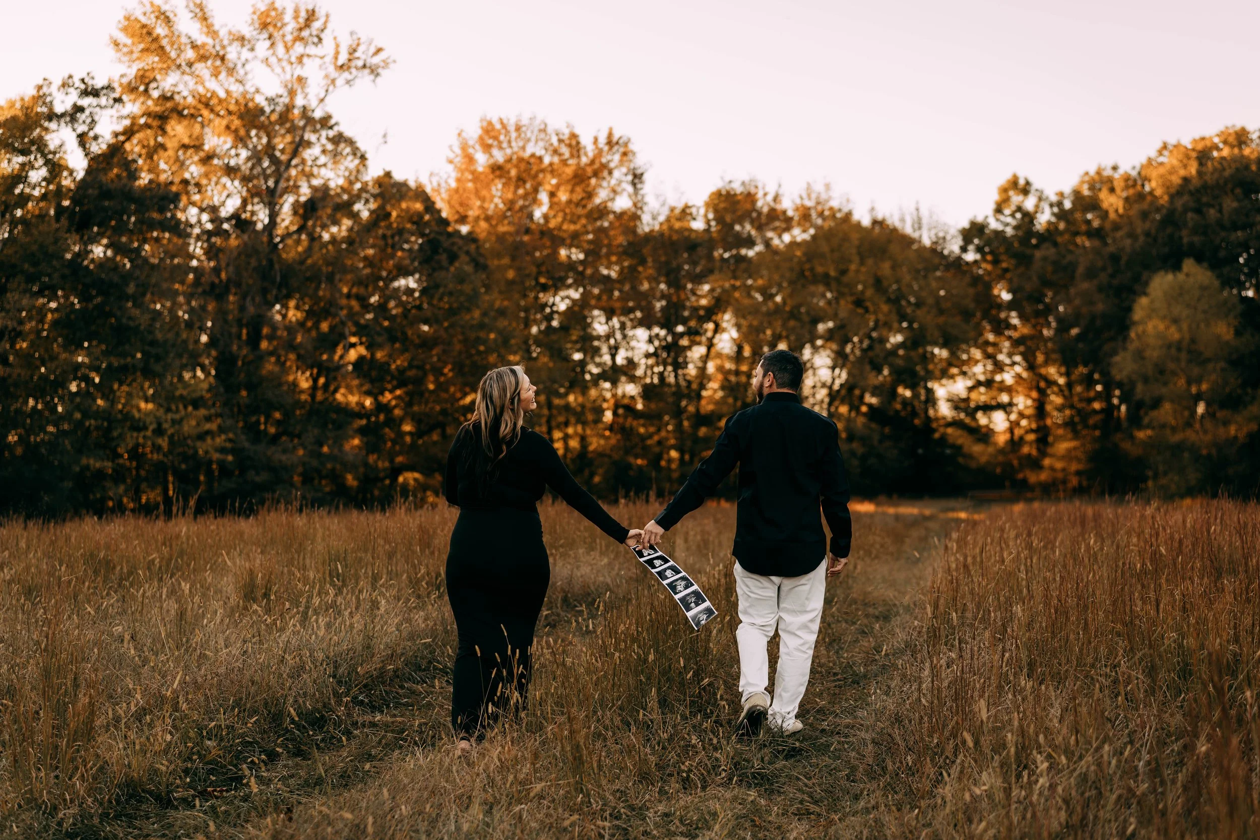 A couple walking through a grassy field at sunset, holding hands, with an ultrasound picture in one person's hand, and trees in the background.