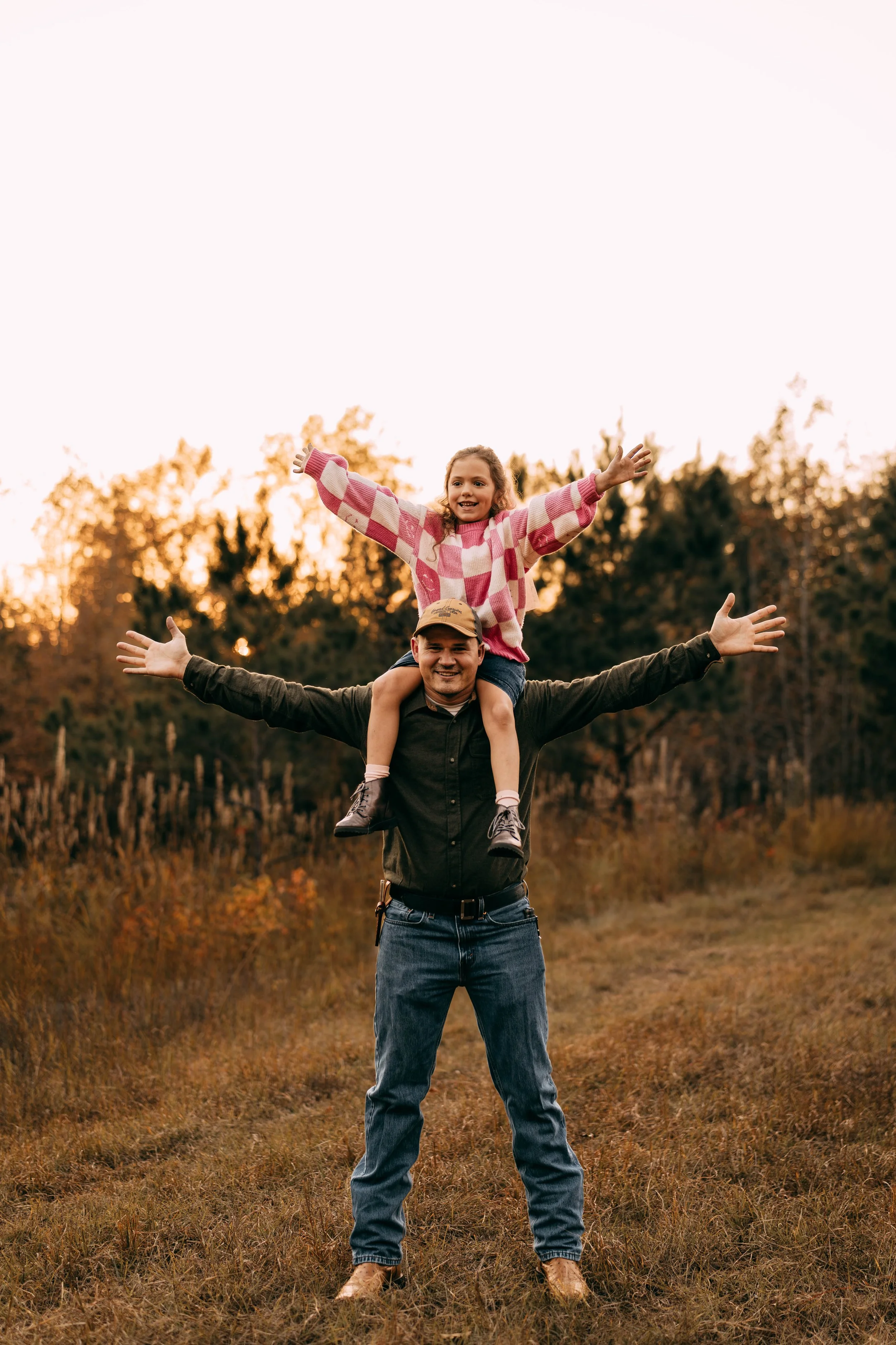 A man giving a piggyback ride to a young girl outdoors during sunset, both smiling with arms outstretched, in a grassy field with trees in the background.