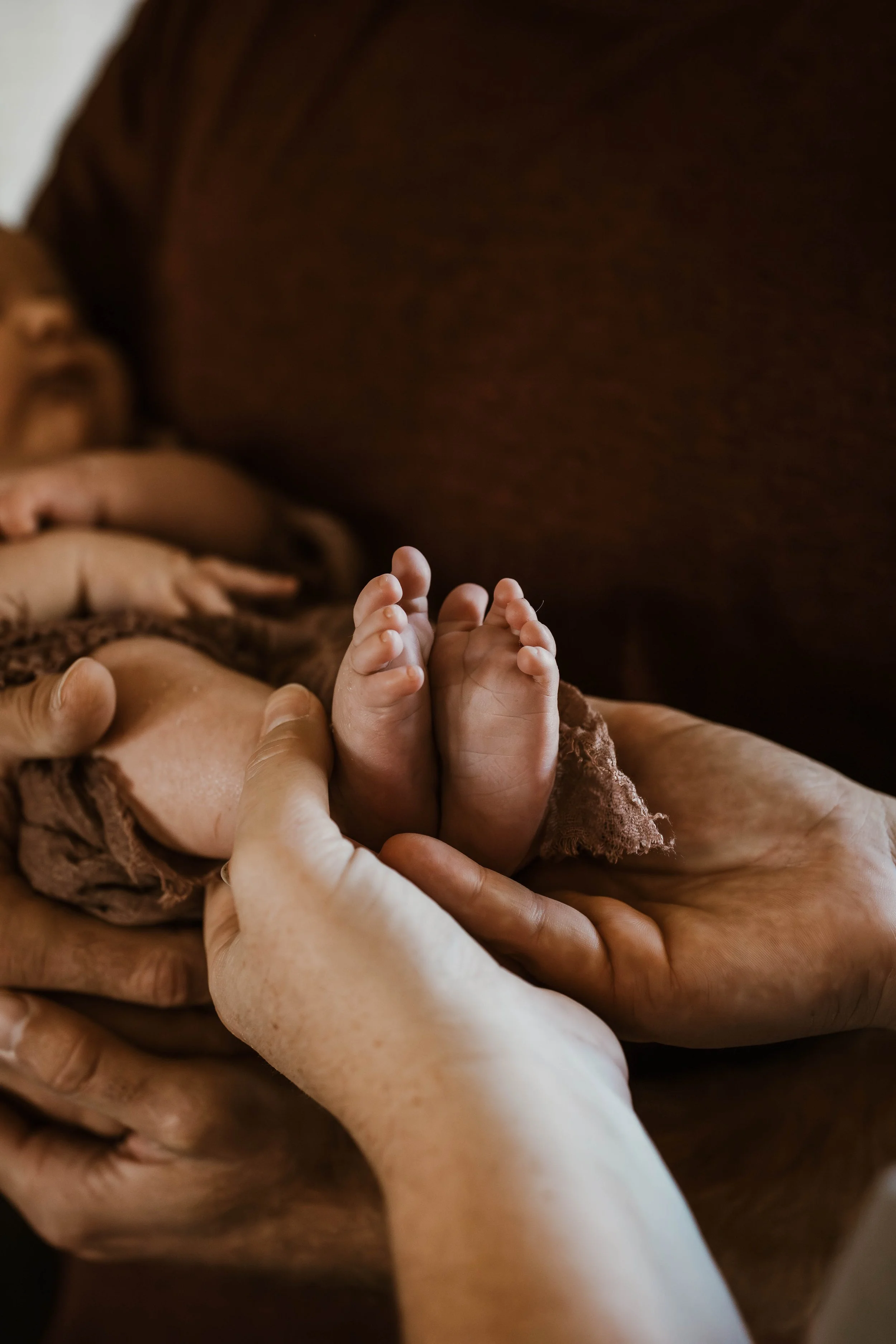 Close-up of a newborn baby's feet being gently held by an adult's hands, with a sleeping baby in the background.