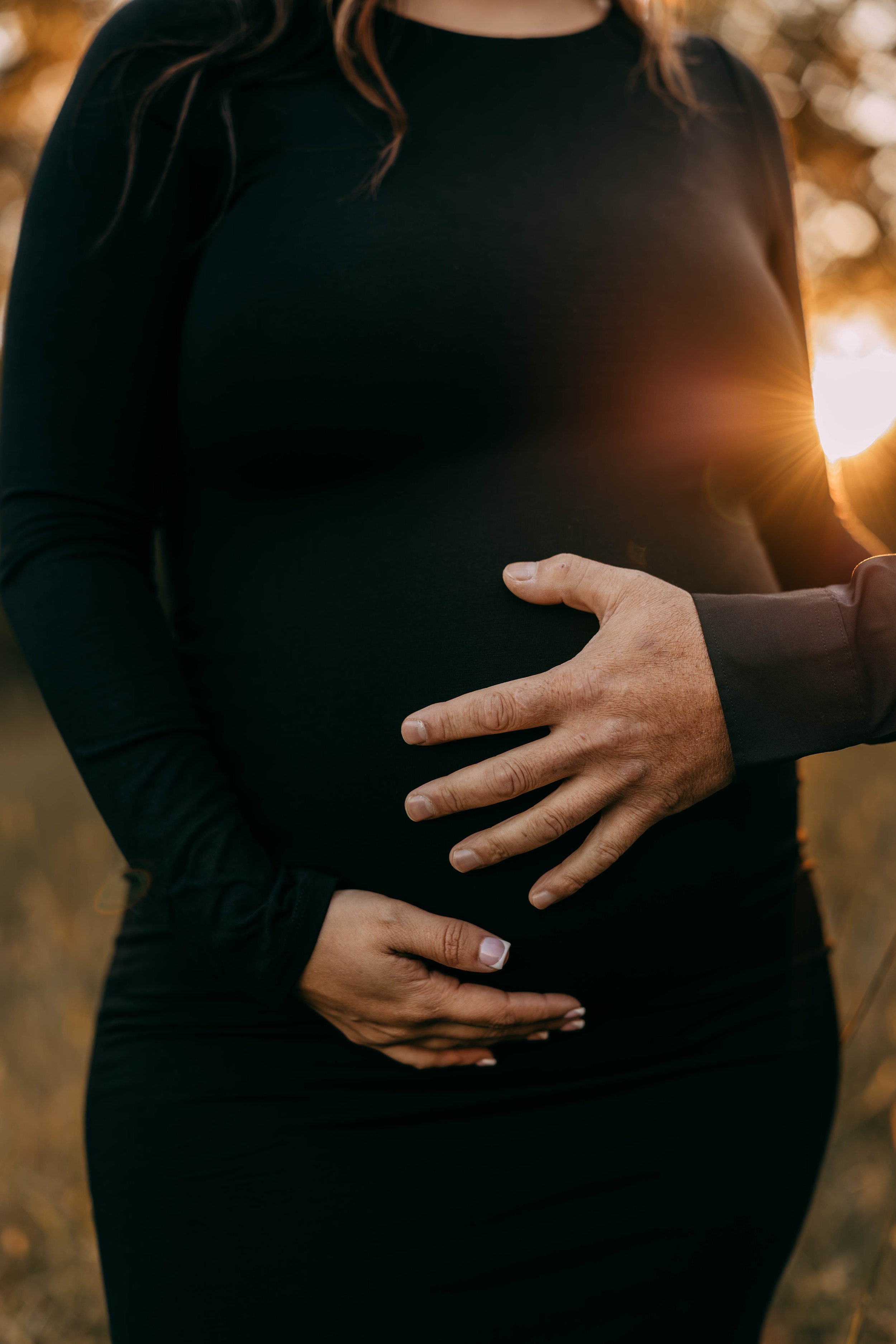 A pregnant woman wearing a black long-sleeve dress, standing outdoors during sunset, with hands gently resting on her belly.