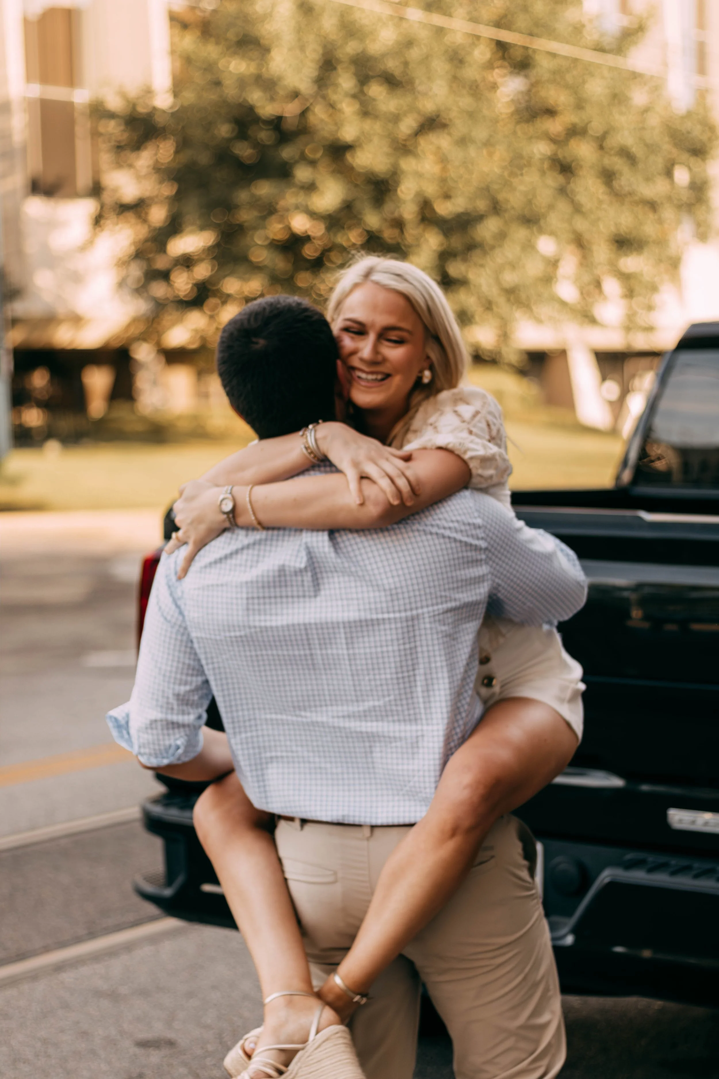 A woman with blonde hair and a man in a checkered shirt embracing outdoors near a black truck, with trees and buildings in the background.