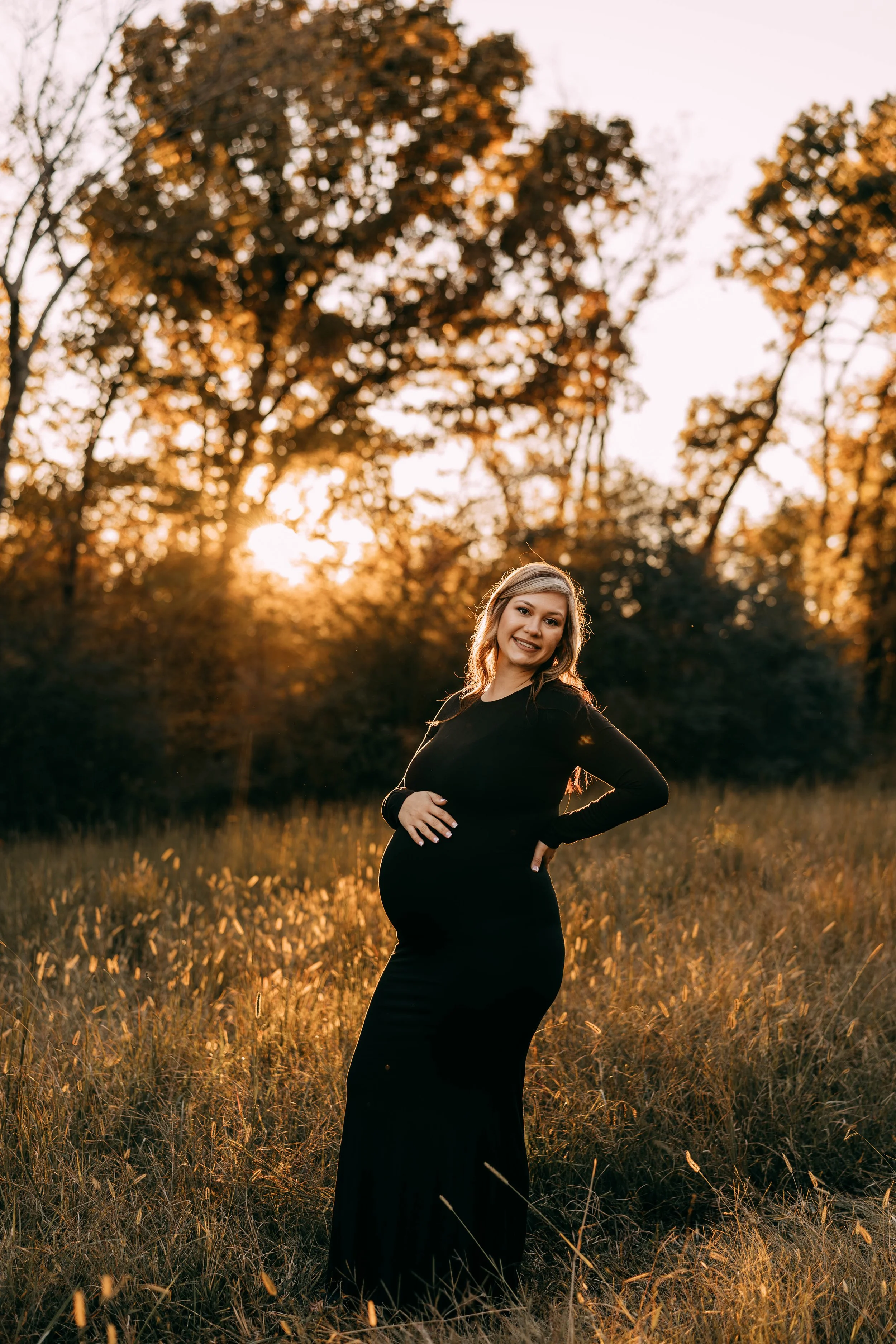 A pregnant woman in a long black dress standing in a field during sunset, smiling at the camera.