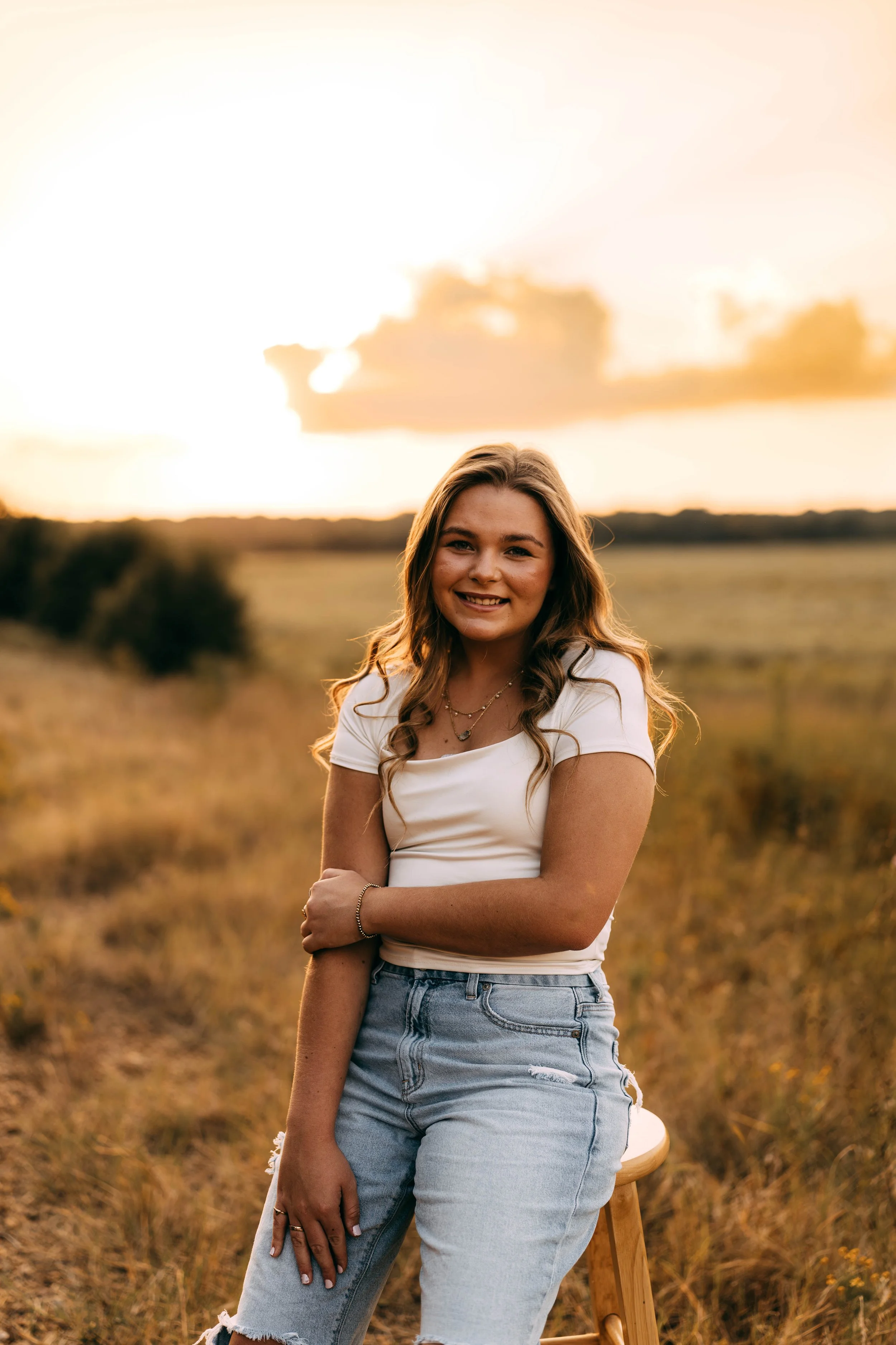 A young woman with wavy brown hair and a white T-shirt, smiling and sitting on a stool in an open field during sunset.