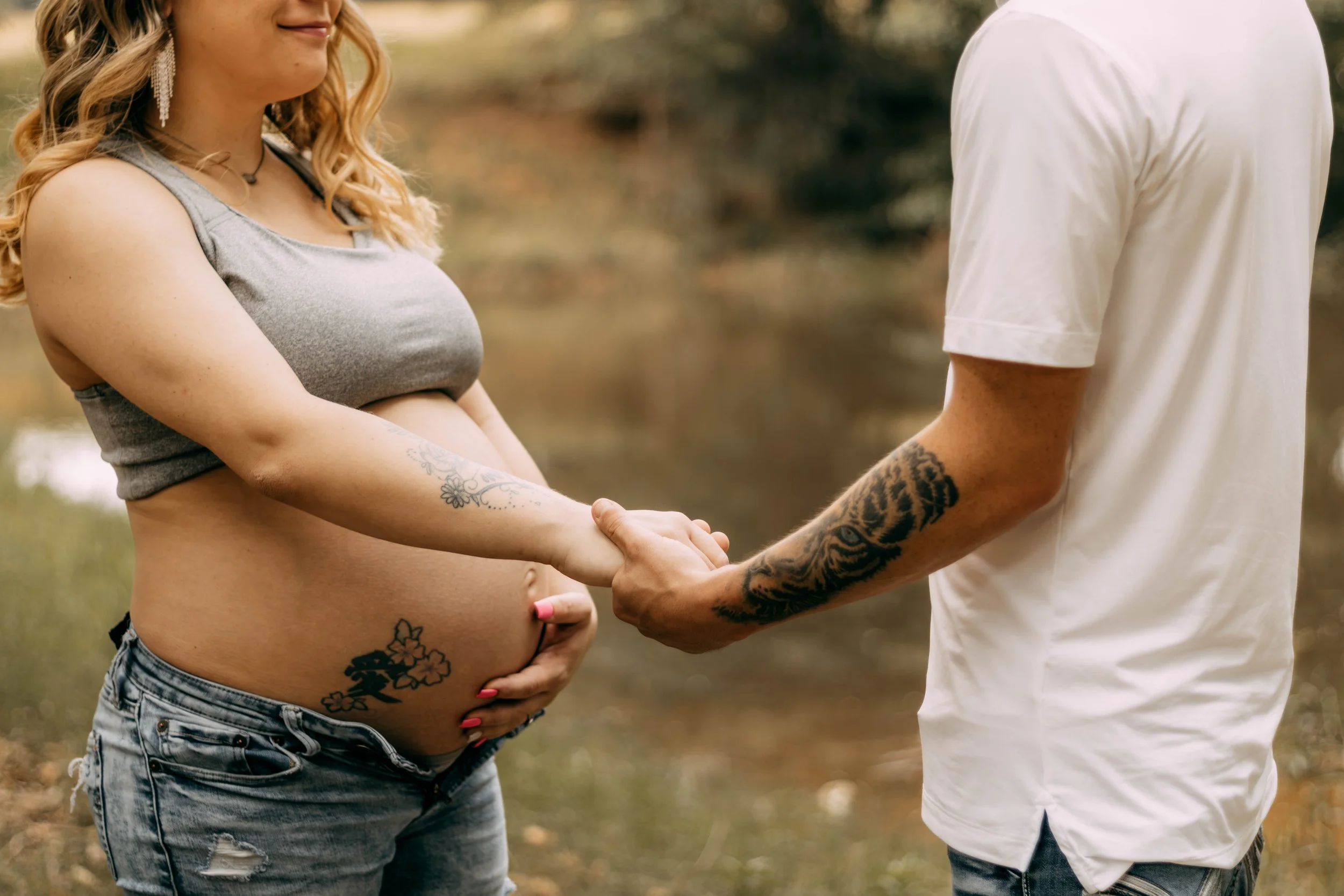 A pregnant woman in a gray top holding her belly and holding hands with a man outdoors near a body of water, both with tattoos.