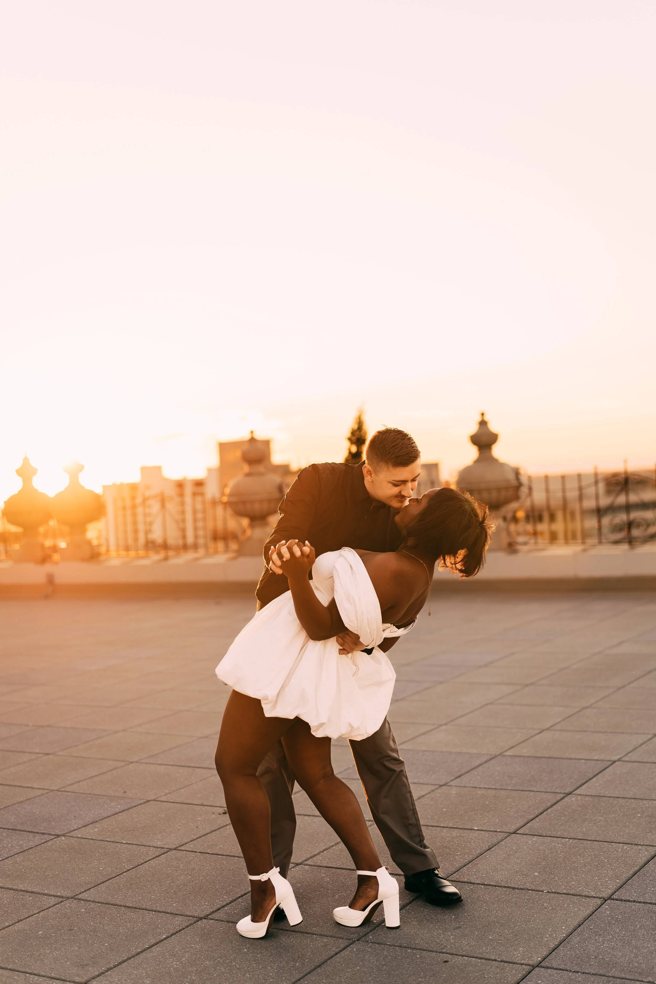 A couple dancing romantically on a rooftop at sunset, with city buildings and decorative structures in the background.