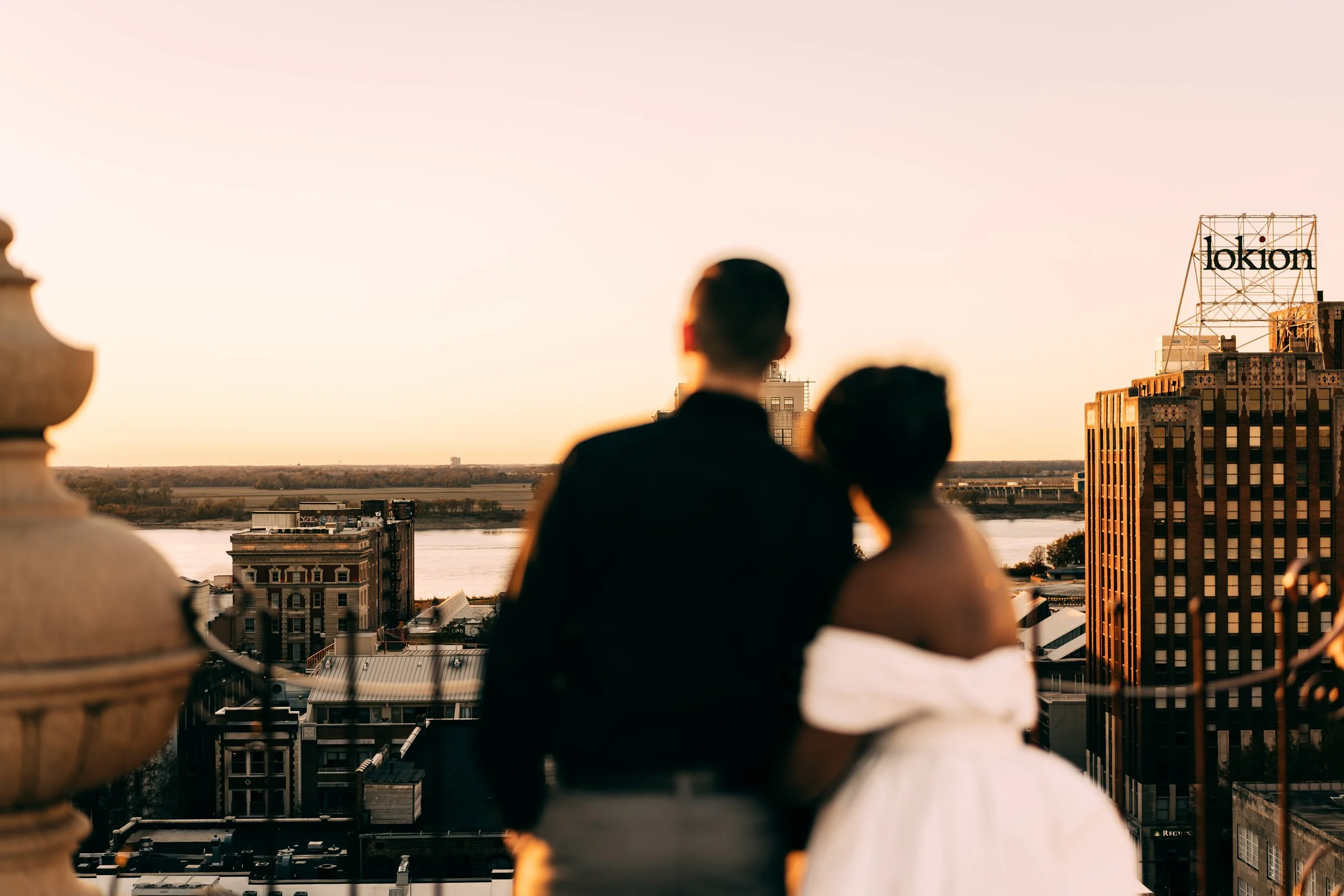 A couple dressed in formal attire standing on a balcony overlooking a cityscape at sunset, with the river and buildings in the background.