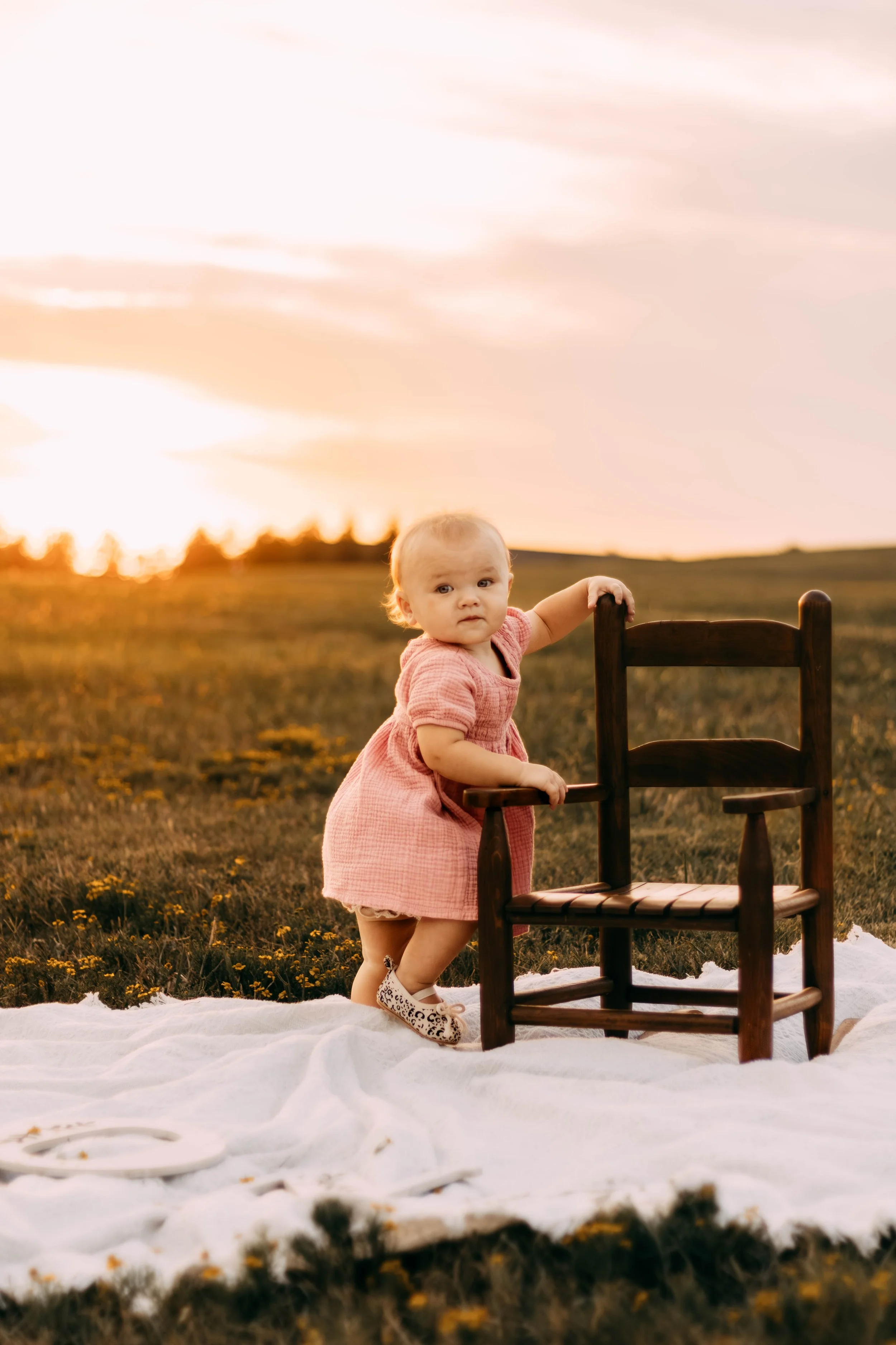 A young girl in a pink dress standing outdoors at sunset, holding onto a small wooden chair on a white blanket with yellow flowers scattered around.