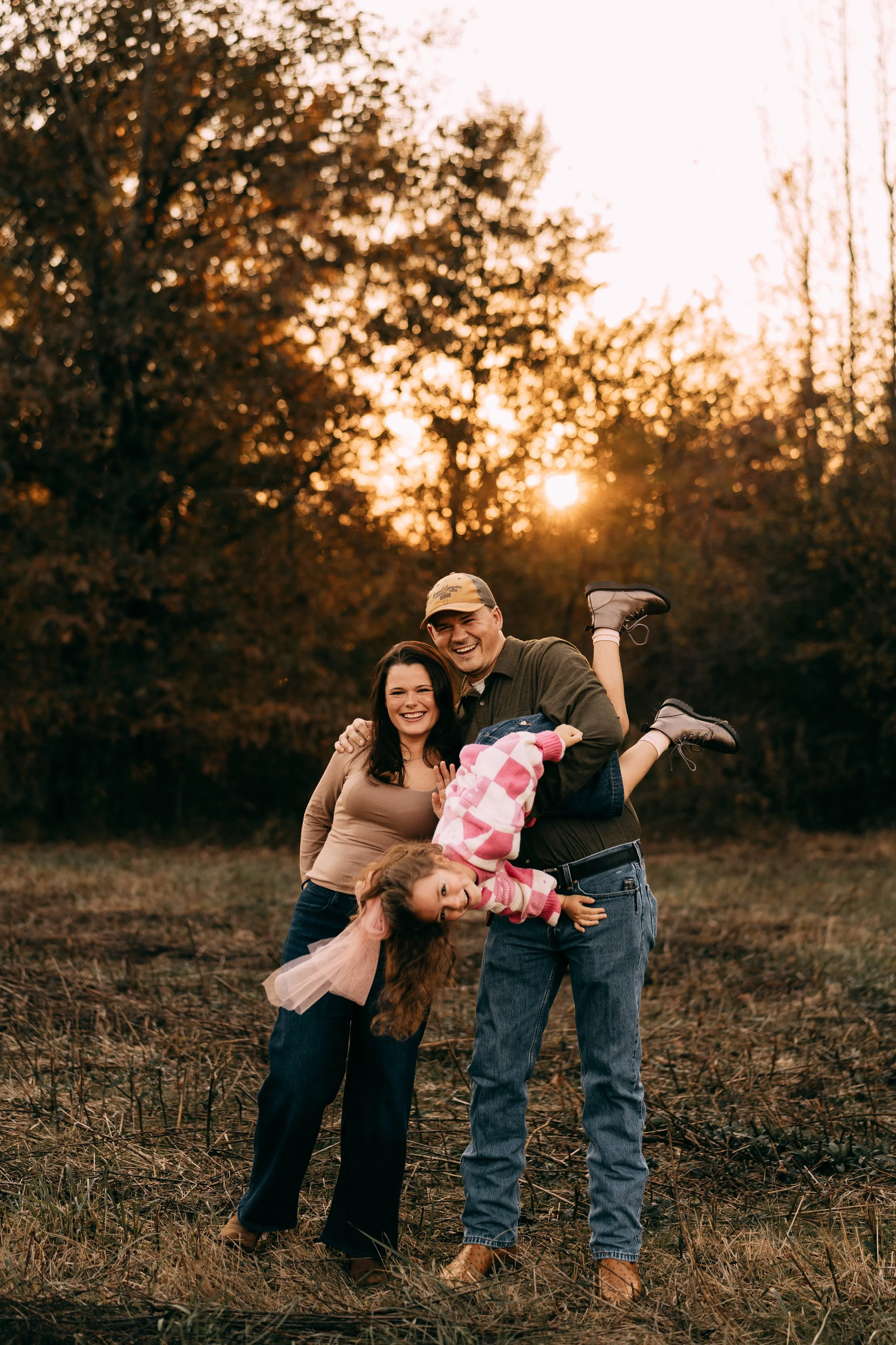 Family of four outdoors during sunset, with two women, one man, and a young girl, playing and smiling together in a field with trees in the background.