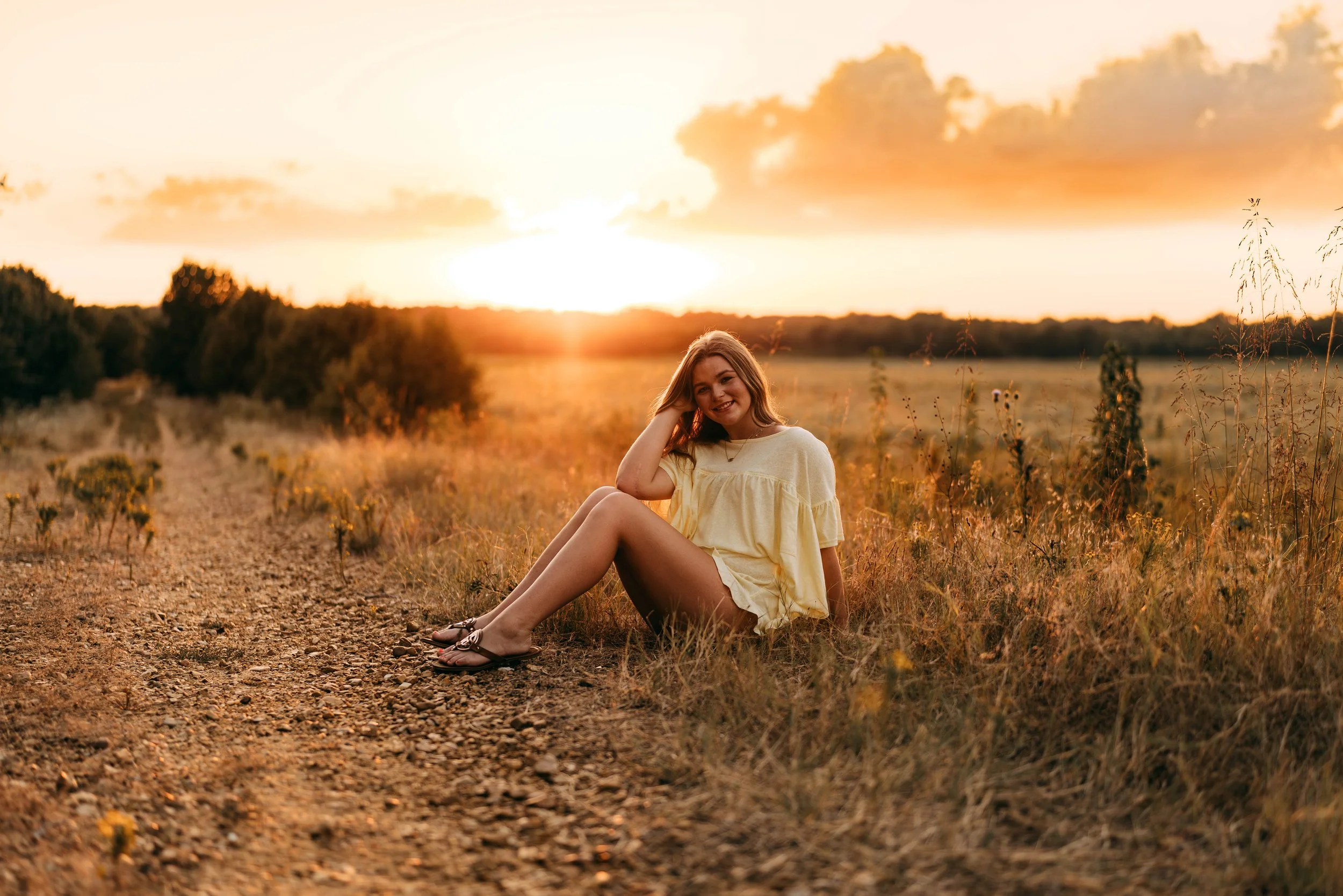 Young woman sitting on a dirt path in a field at sunset, smiling and wearing a yellow dress.