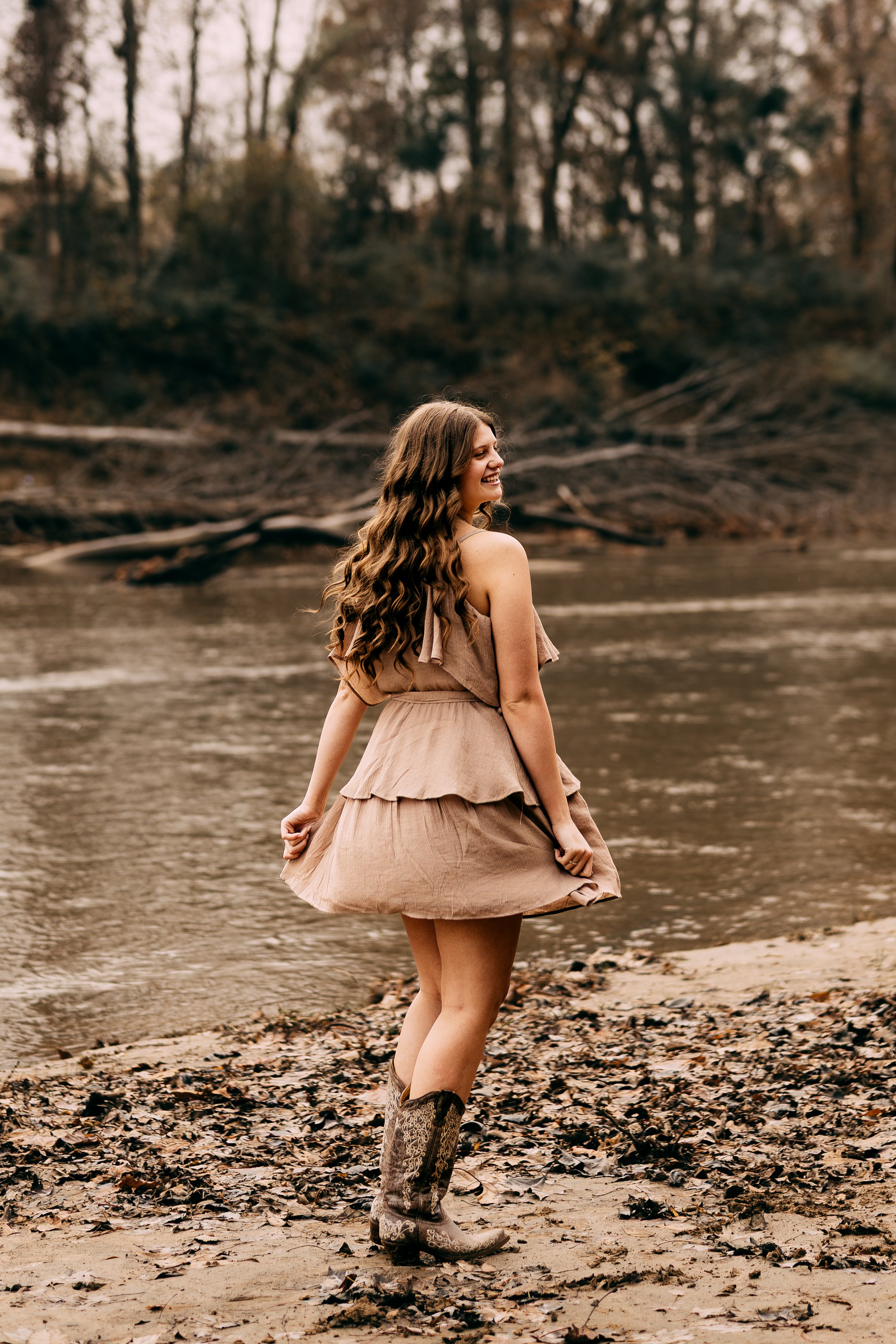 A woman with long, wavy hair wearing a beige dress and cowboy boots standing on a riverbank, smiling and looking to the side with trees in the background.