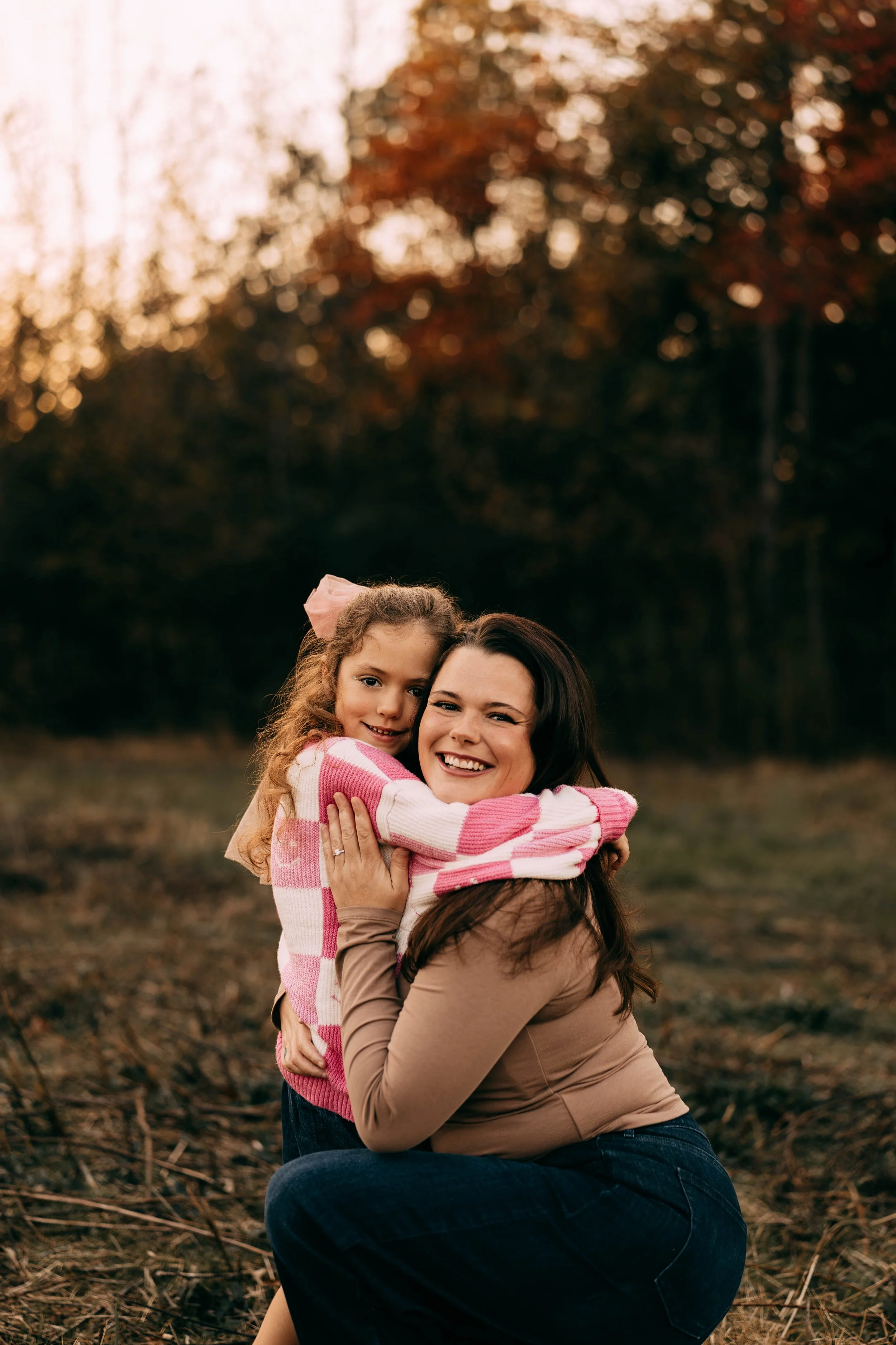 A woman and a young girl hugging outdoors during sunset, with autumn trees in the background.