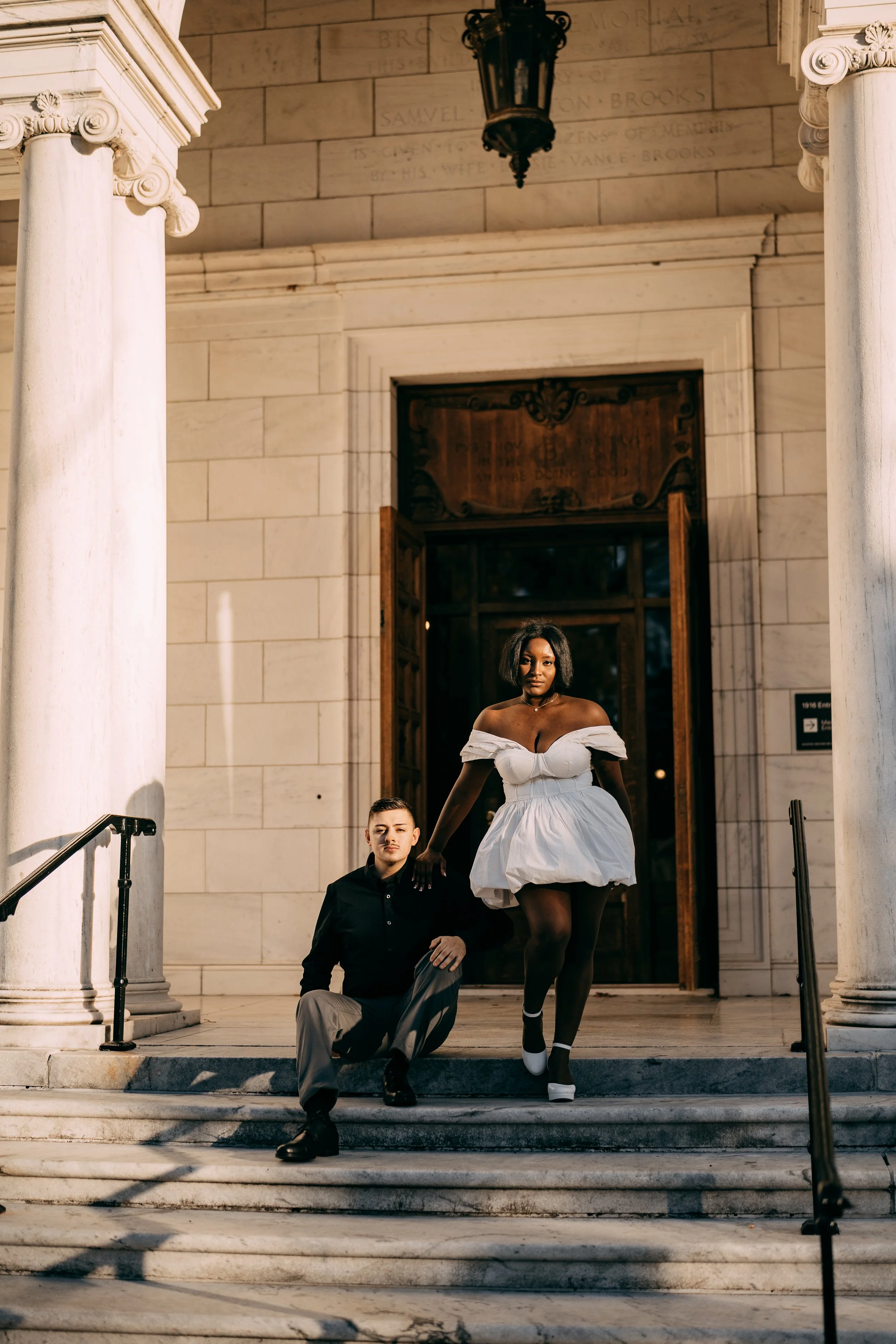 A woman in a white off-shoulder dress, standing on steps with a man kneeling beside her, in front of a historic building with columns and a lantern hanging above.