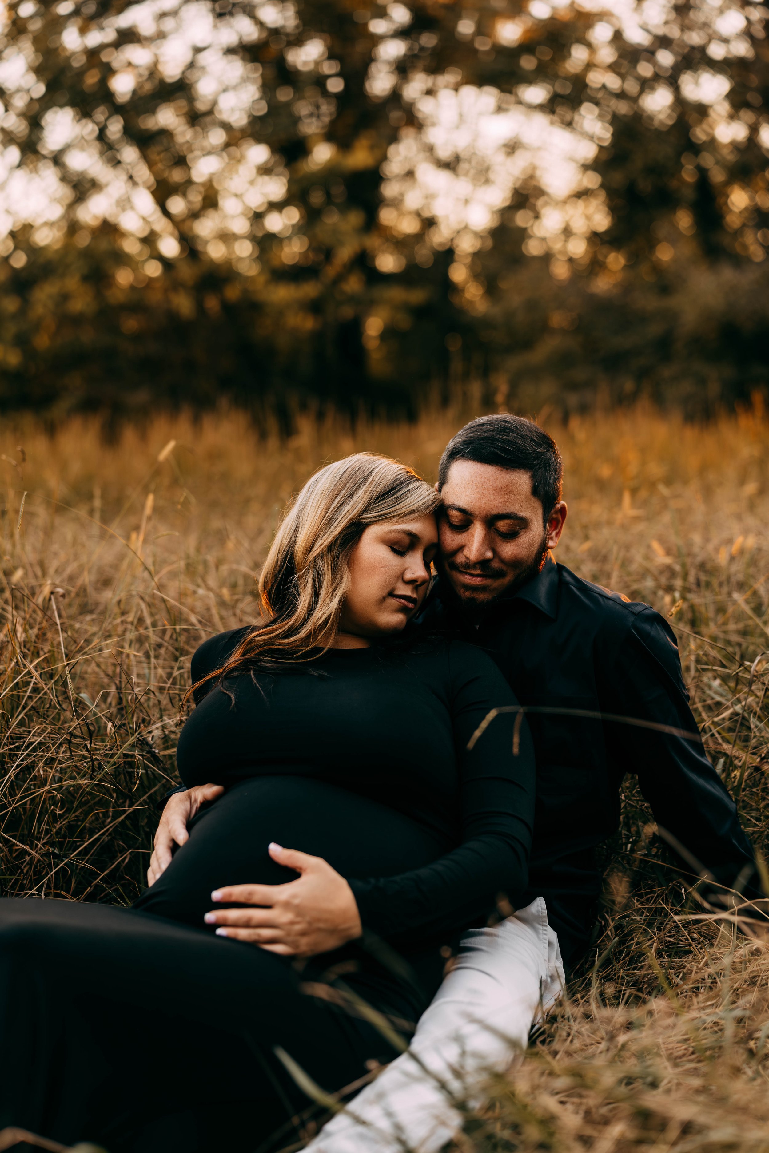 A couple sitting in a field during sunset, with the woman visibly pregnant, embracing affectionately with closed eyes.
