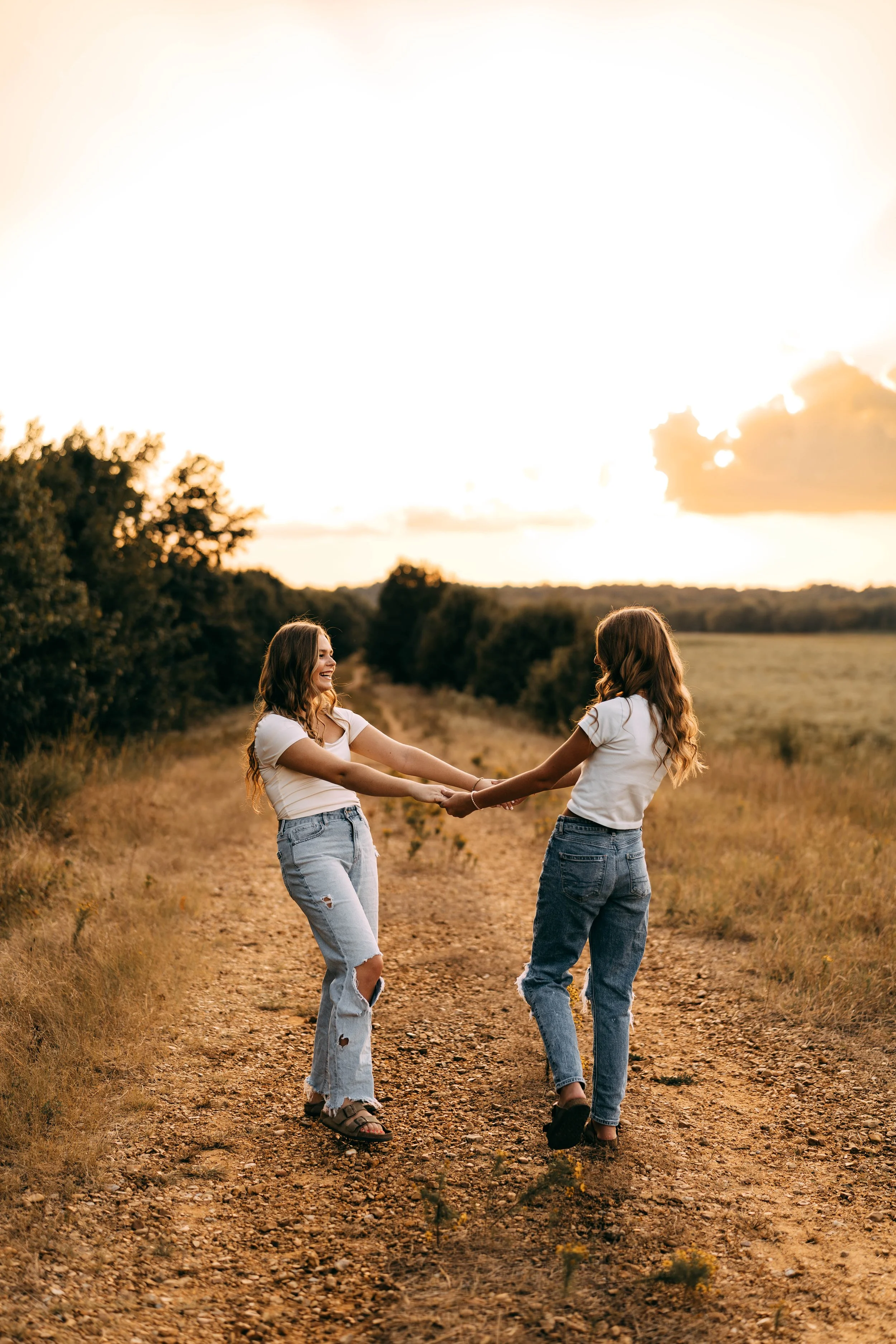 Two young women holding hands and laughing while standing on a dirt path in a field at sunset.