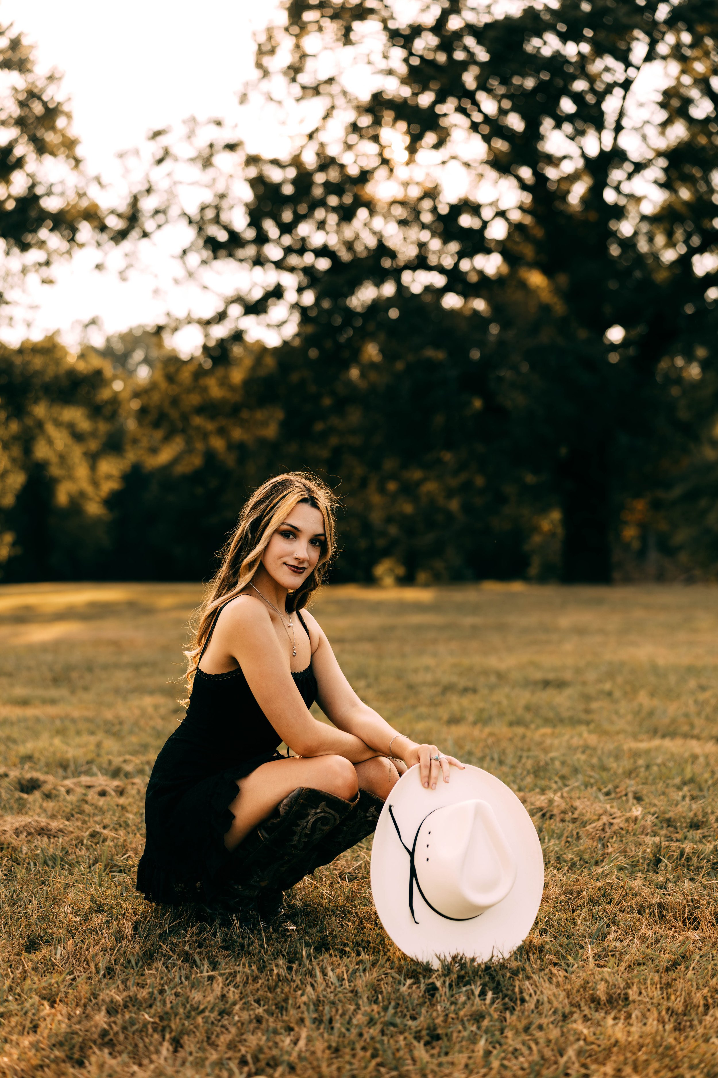 A young woman in a black dress is kneeling on grass in a park during sunset, holding a white hat with a black ribbon.