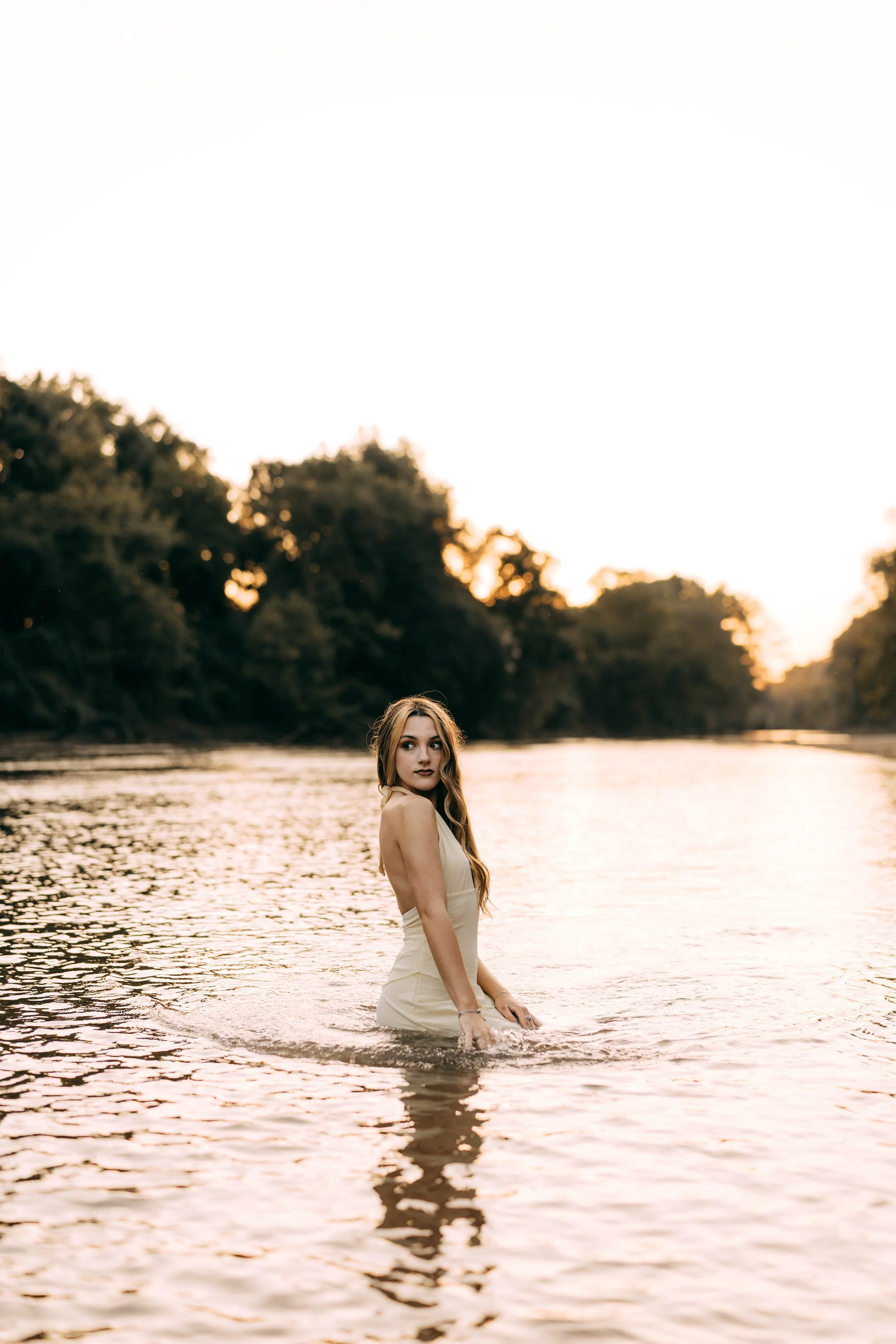 A woman in a cream-colored dress standing waist-deep in a river during sunset, with trees in the background.
