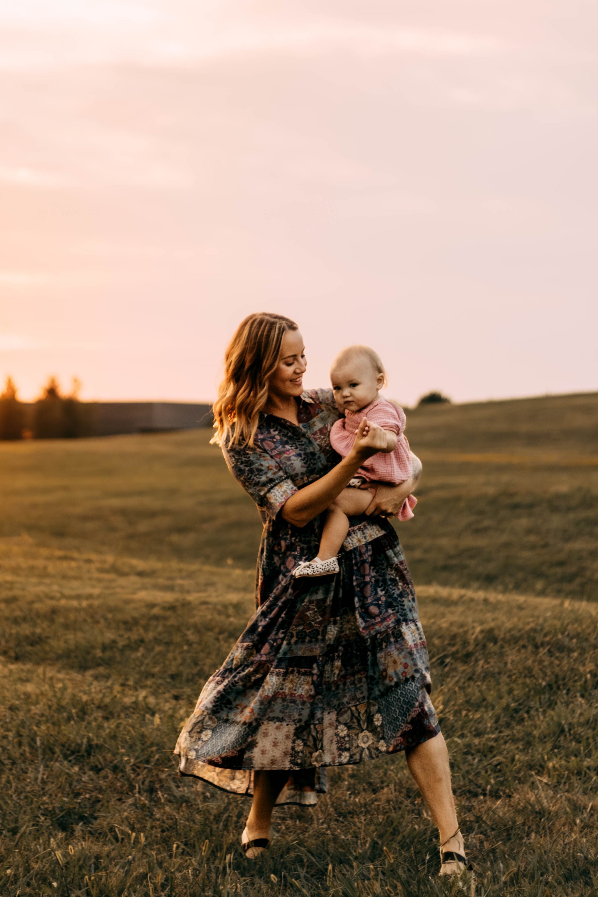 A woman holding a young girl in a grassy field during sunset, both smiling.