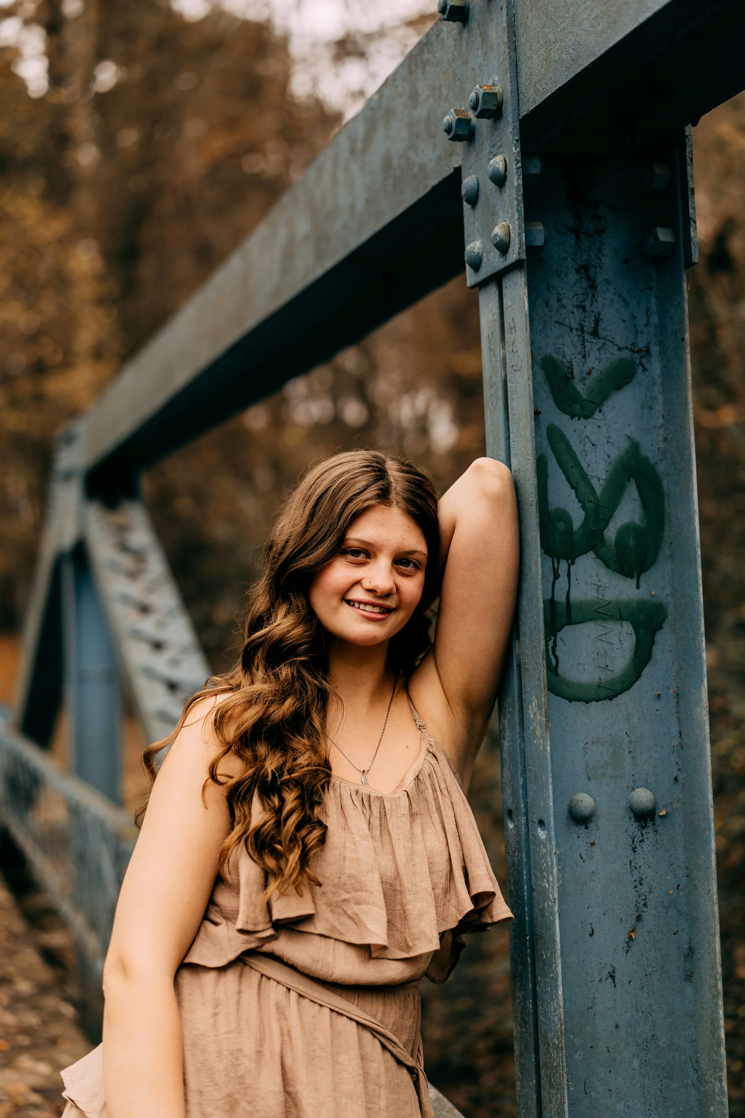 A young woman with long, wavy brown hair and a tan dress smiling and leaning against a blue metal structure with graffiti, outdoors during autumn with blurred fall trees in the background.
