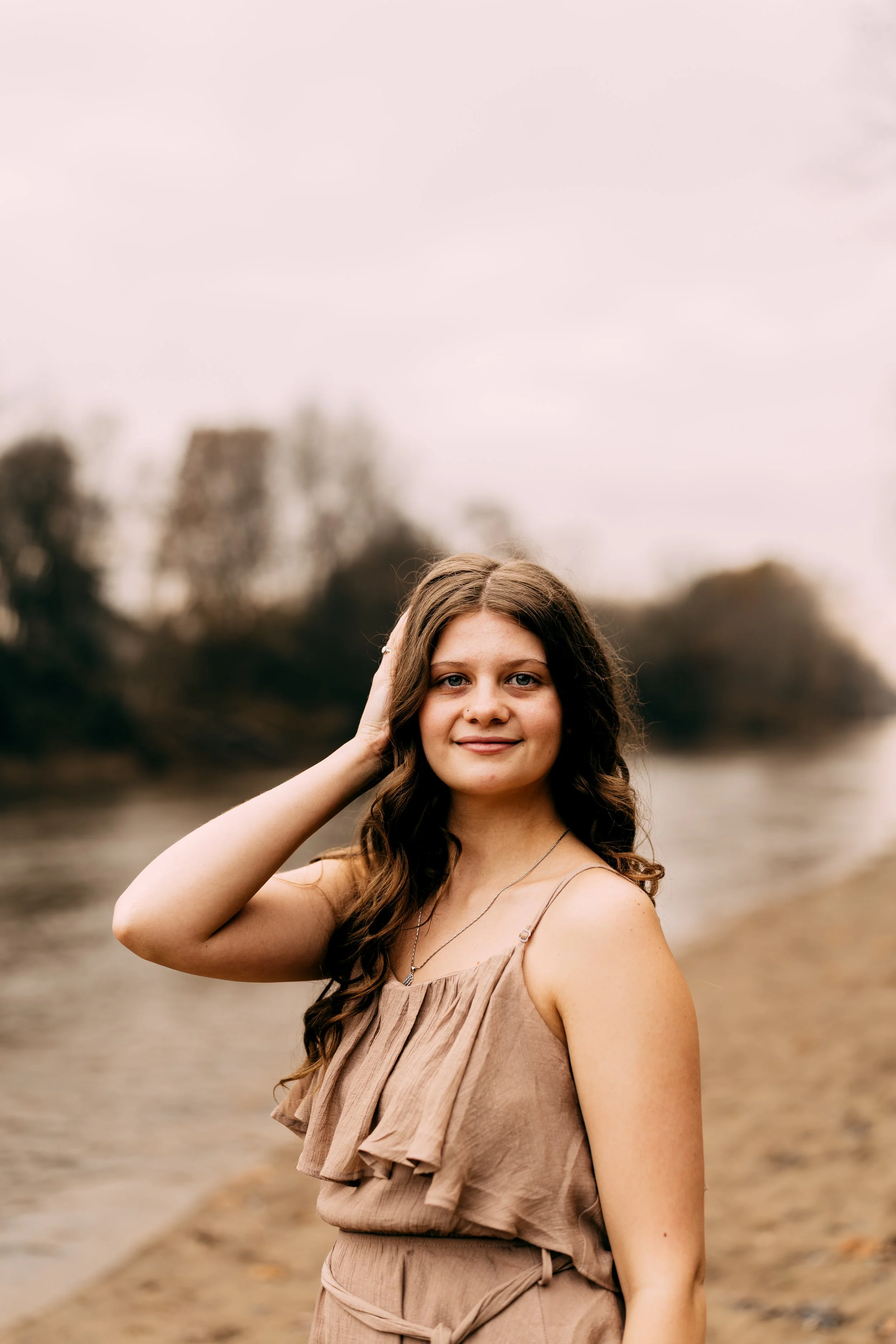 A young woman with long brown hair standing near a river on an overcast day.