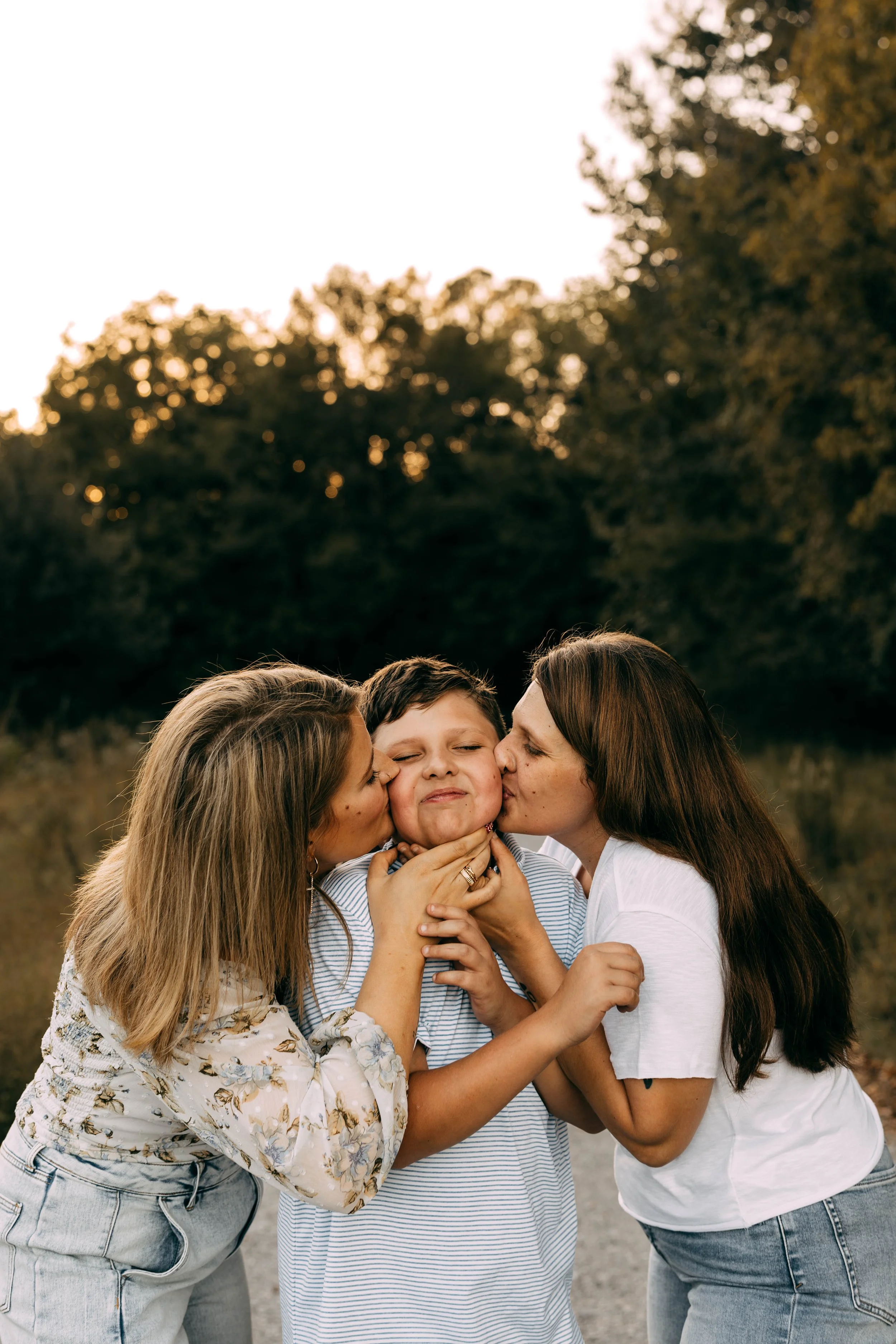 Two women kissing a young boy on each cheek in an outdoor setting during sunset.