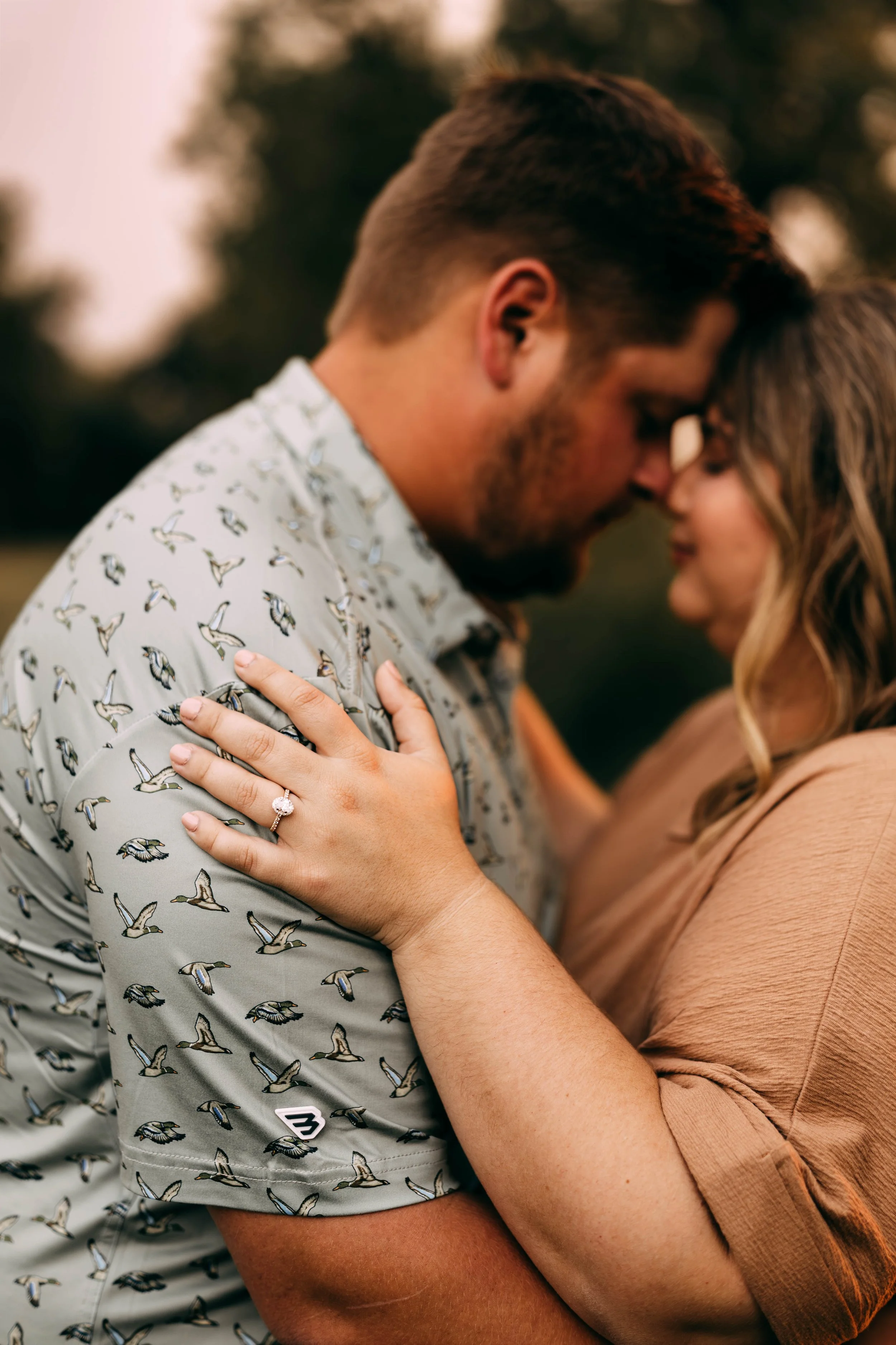 A couple close together, foreheads touching outdoors during sunset. The woman, with a visible engagement ring, has her hand on the man's shoulder, and they are about to kiss.