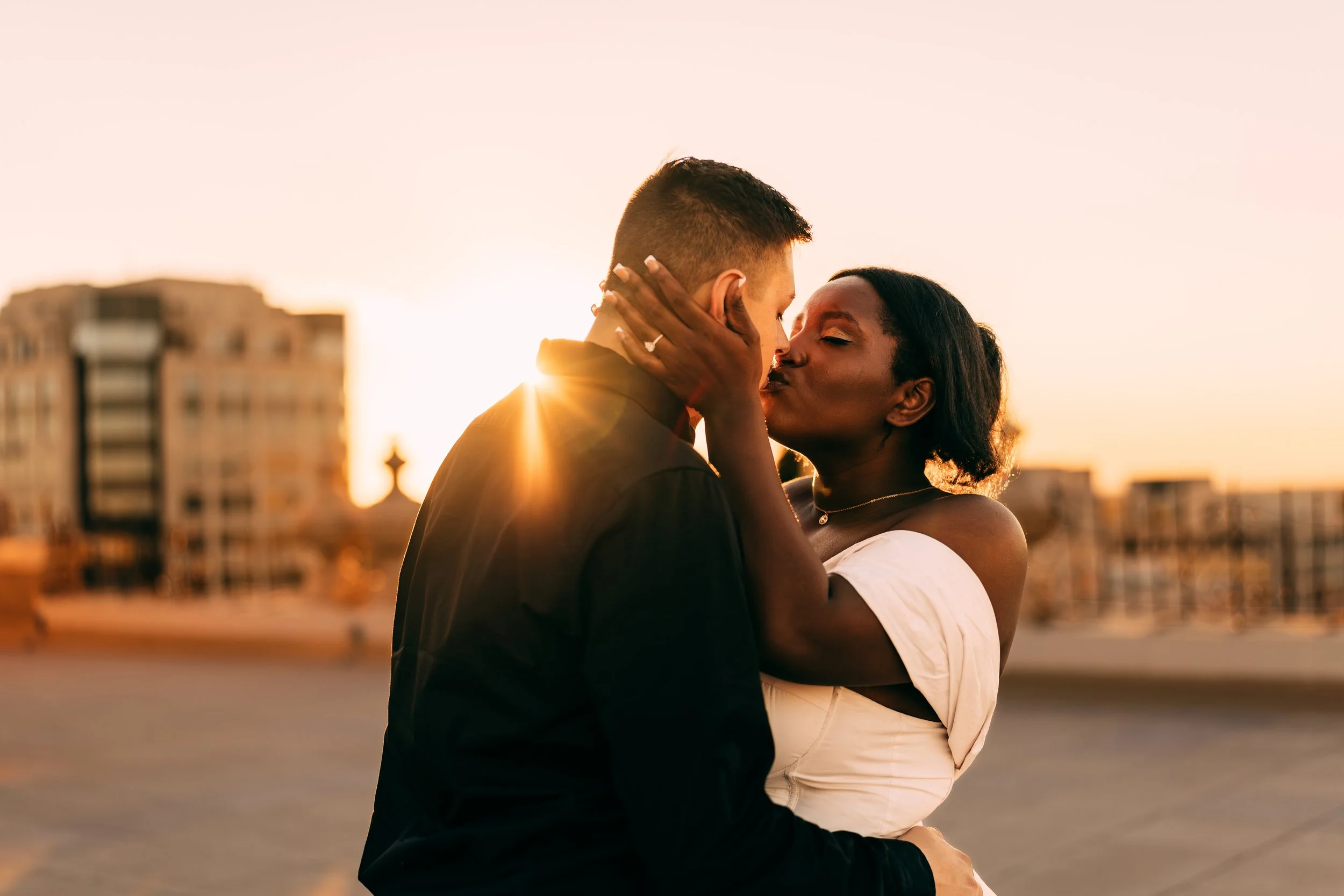 A couple sharing a kiss at sunset on a city rooftop, with buildings in the background.