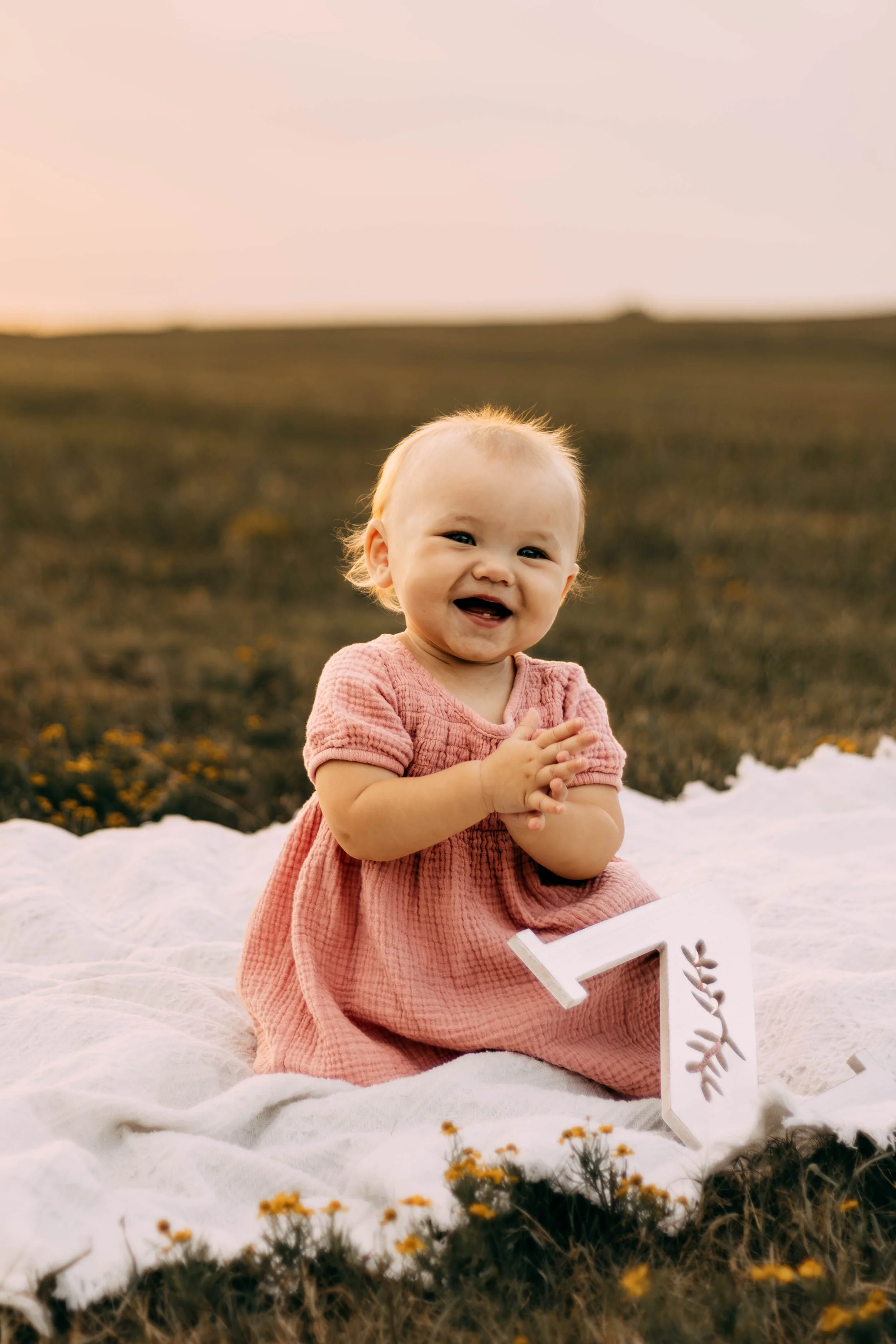 A smiling baby girl in a pink dress sits on a white blanket outdoors during sunset, surrounded by small yellow flowers.