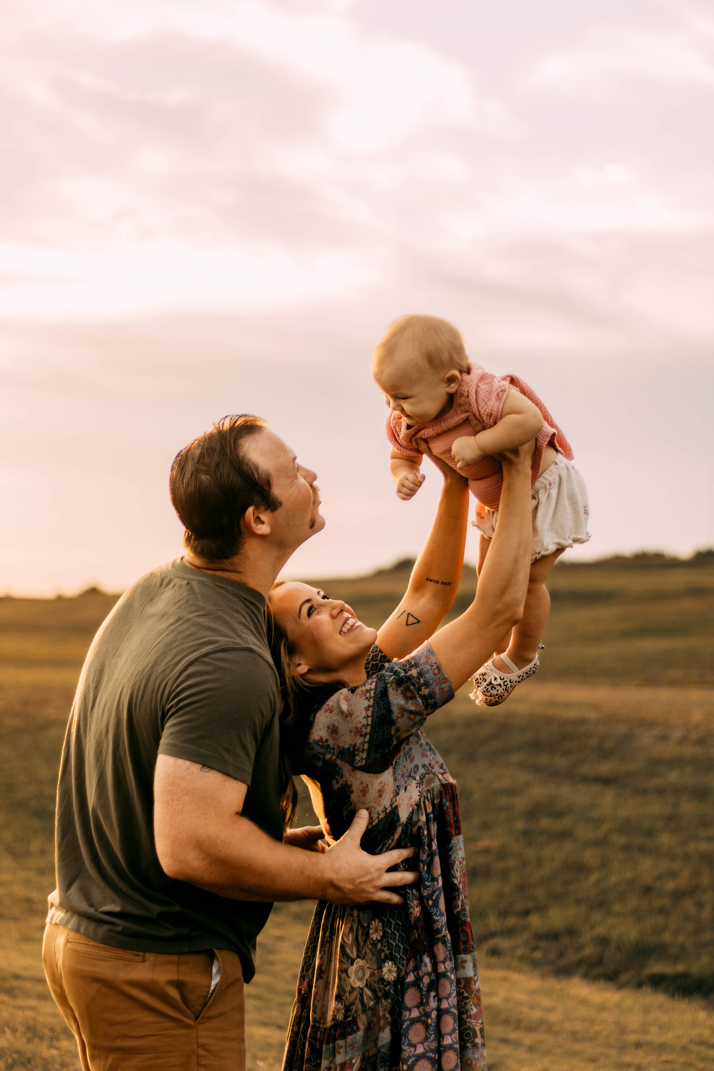 A family outdoors at sunset, with the mother holding up a baby girl while the father stands beside them, smiling and looking at the baby.
