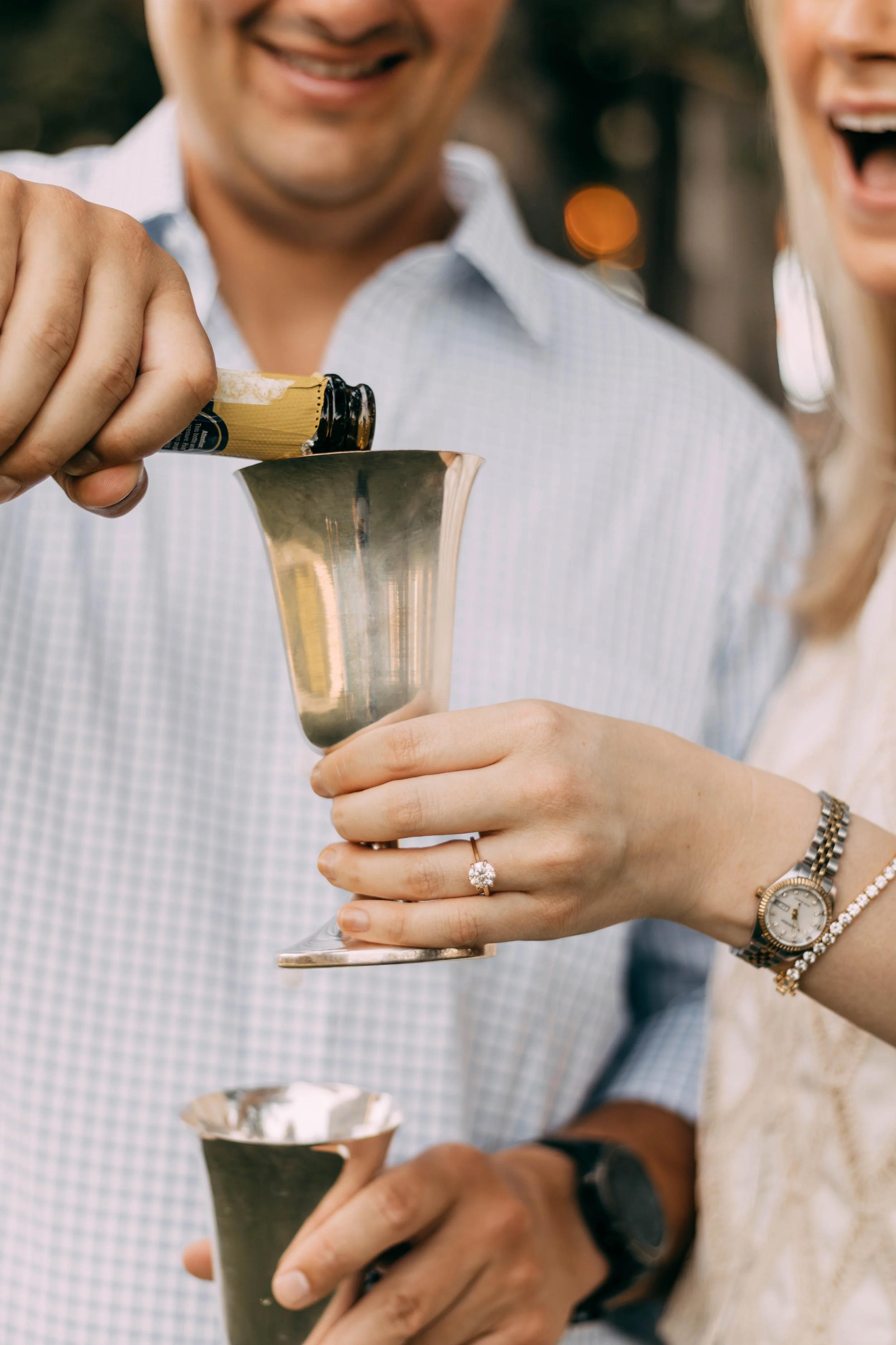 Two people celebrating with a toast, pouring champagne into a metallic cup outdoors, with visible smiling faces and and a woman's hand with a ring and watch.