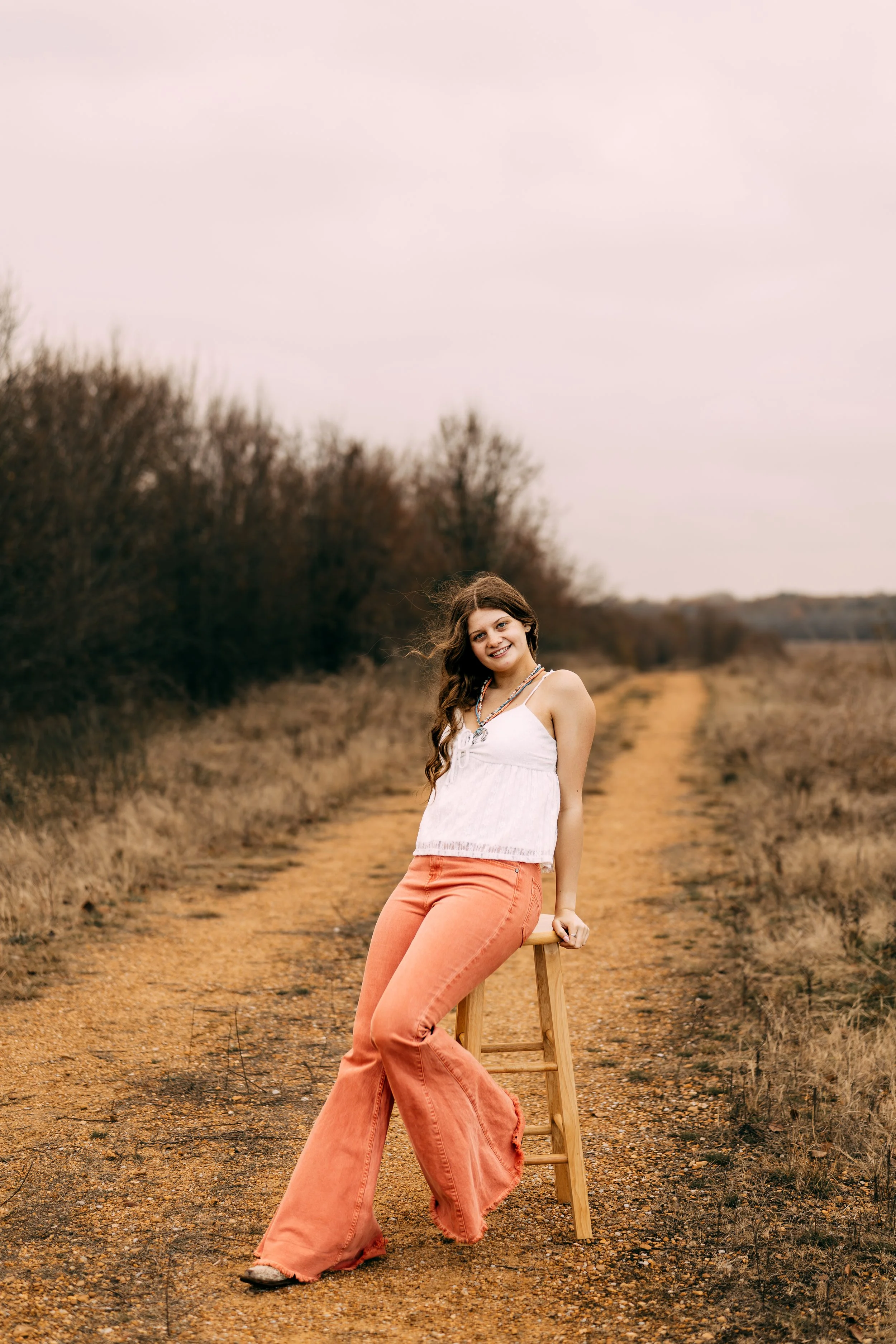 A young woman with long wavy brown hair sitting on a wooden stool on a dirt path in an open field, wearing a white sleeveless top and orange bell-bottom pants, smiling at the camera with a background of leafless trees and a pinkish sky.