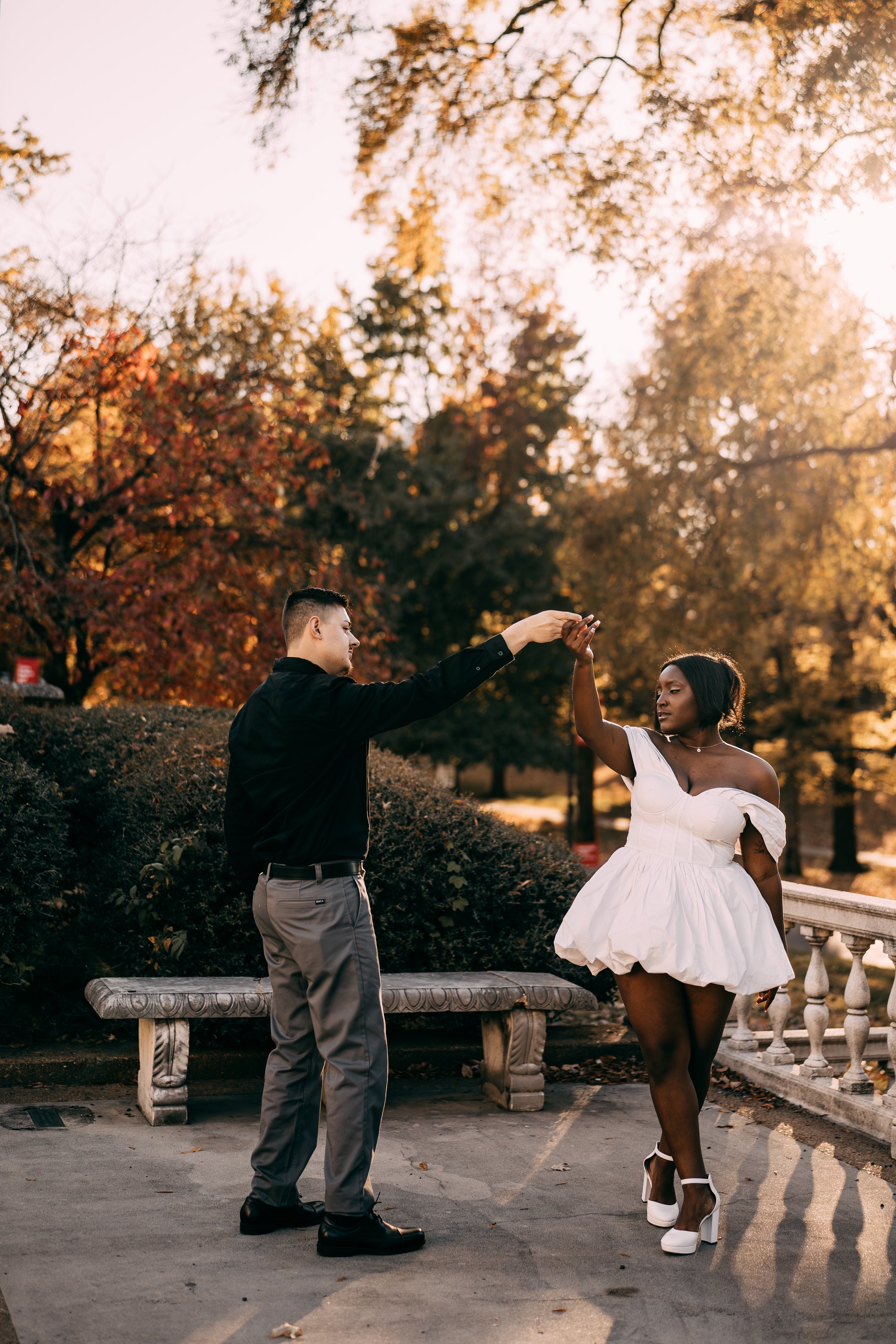 A man and woman dancing outdoors in a park during fall, with colorful autumn trees in the background. The man is holding the woman's hand as she spins around, wearing a white dress and high heels.