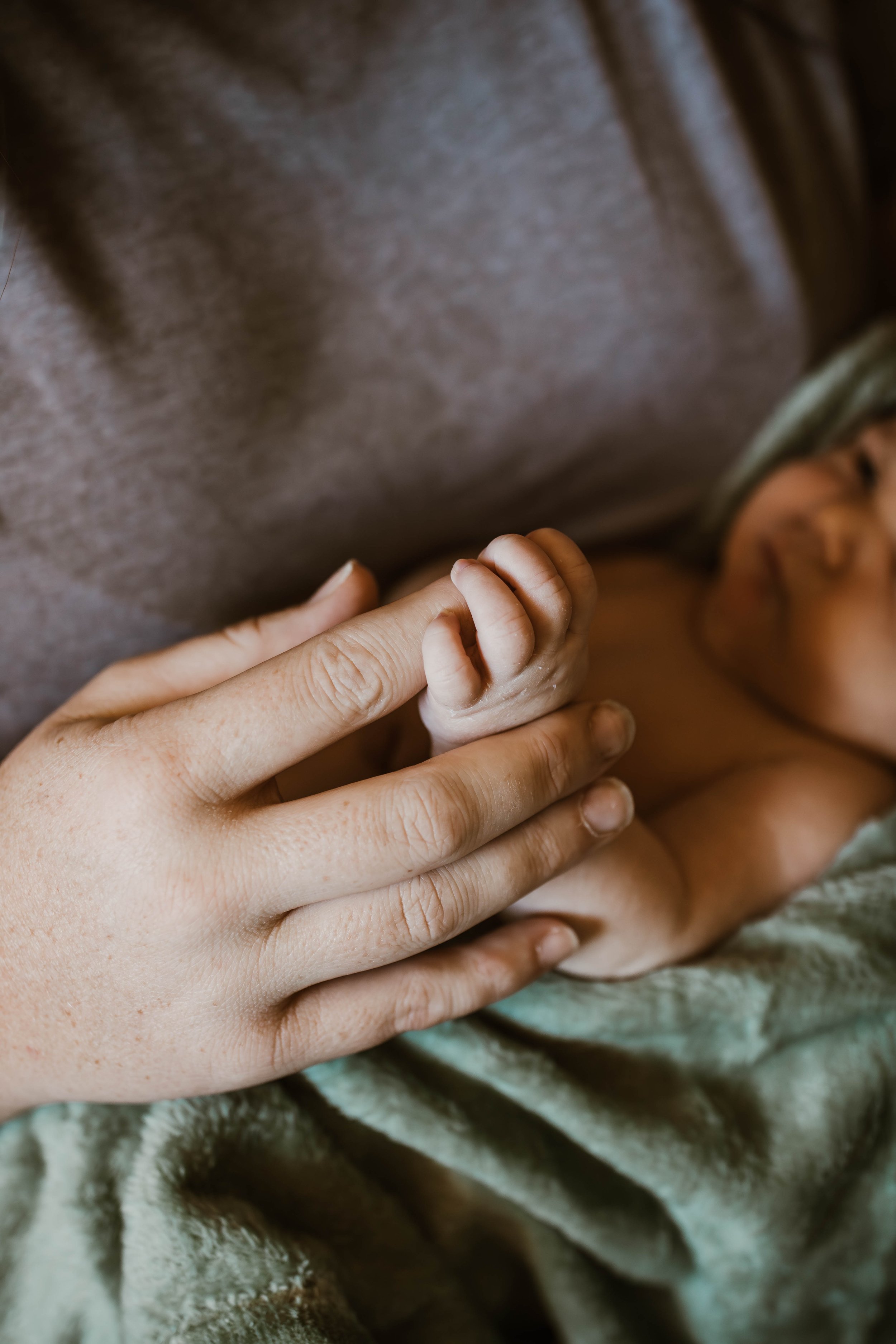 A close-up of a tiny newborn baby holding an adult's finger, lying on a soft blanket with part of their face slightly visible in the background.