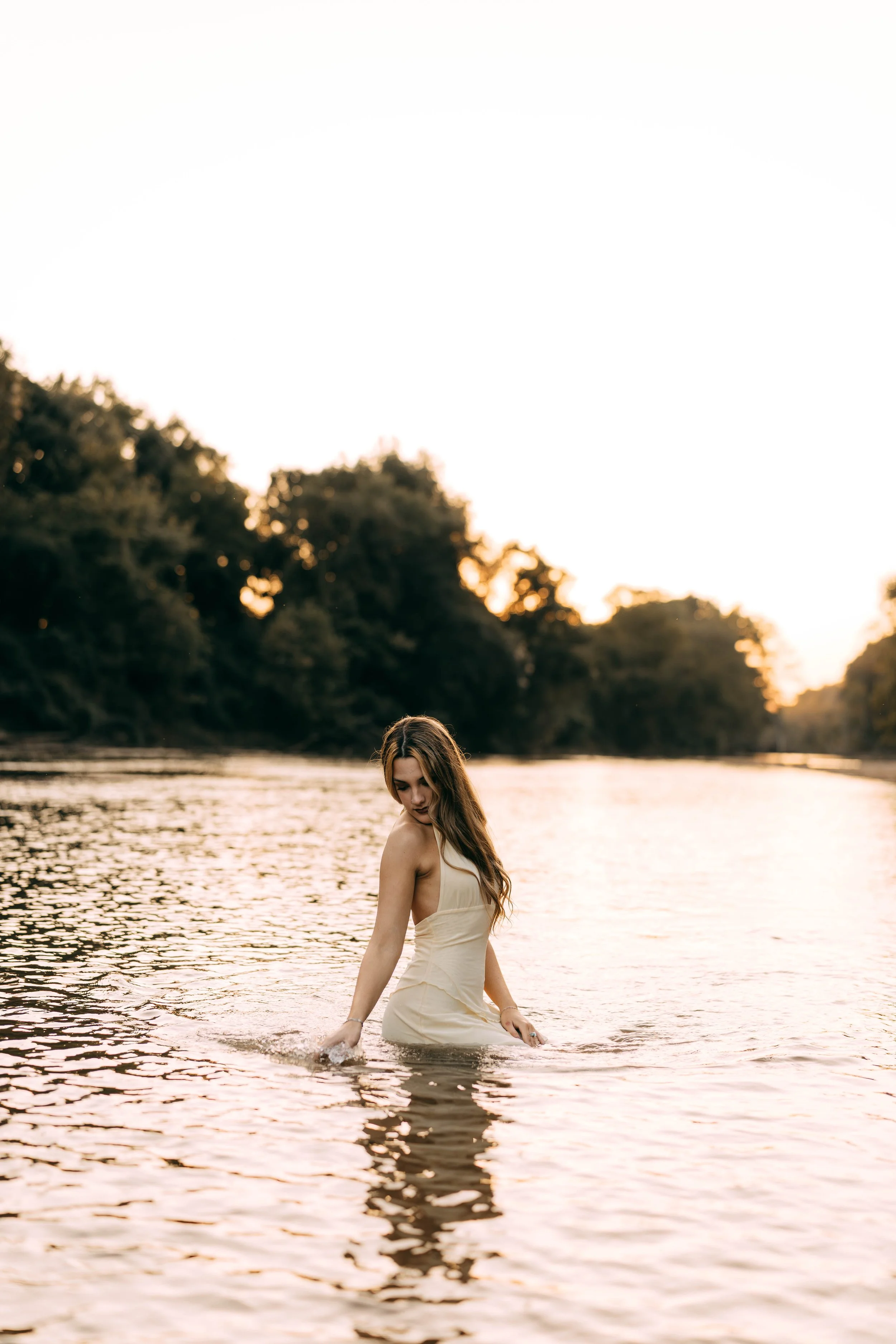 A young woman in a beige dress standing in a calm river at sunset, with trees in the background and soft lighting.