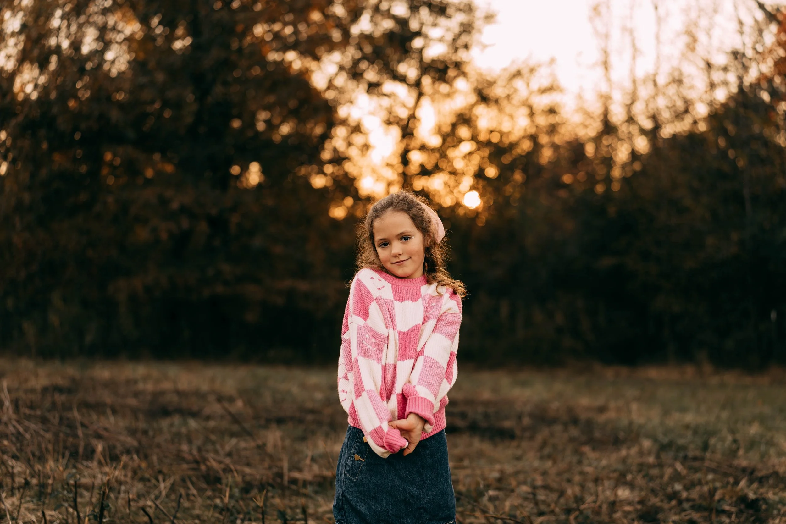 A young girl standing outdoors in a field at sunset, wearing a pink and white sweater and dark skirt, with trees in the background.