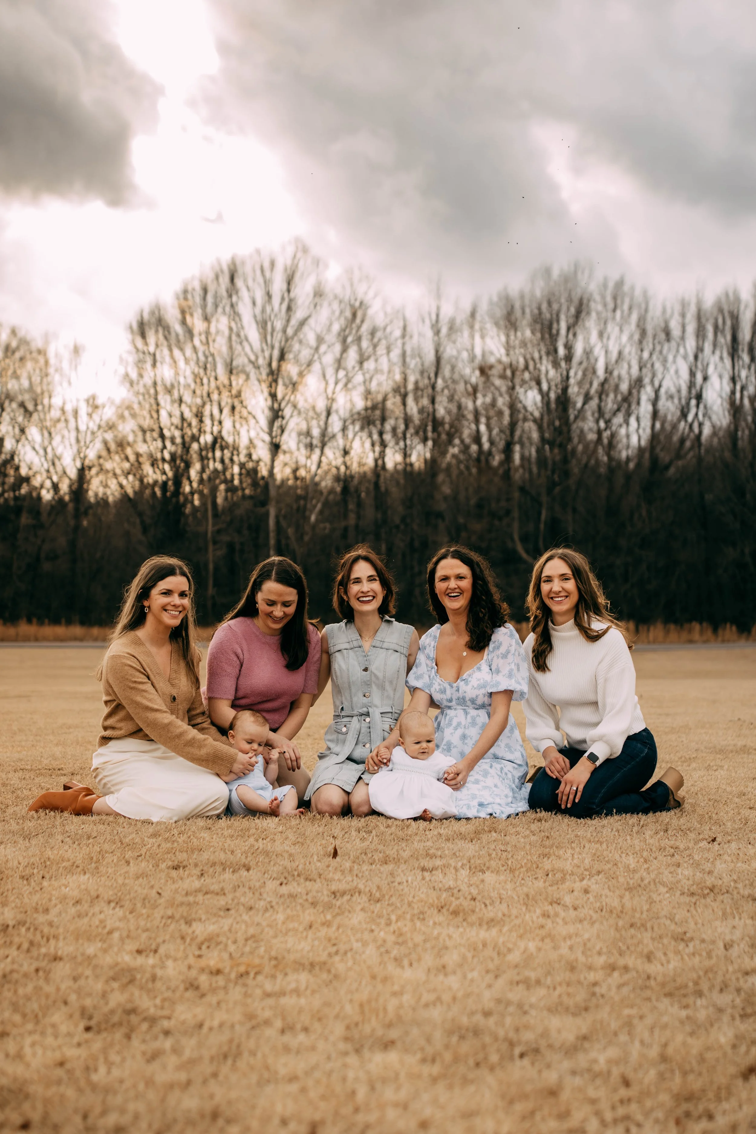 A group of six women, including two babies, sitting on a grassy field outdoors with leafless trees in the background and a cloudy sky.