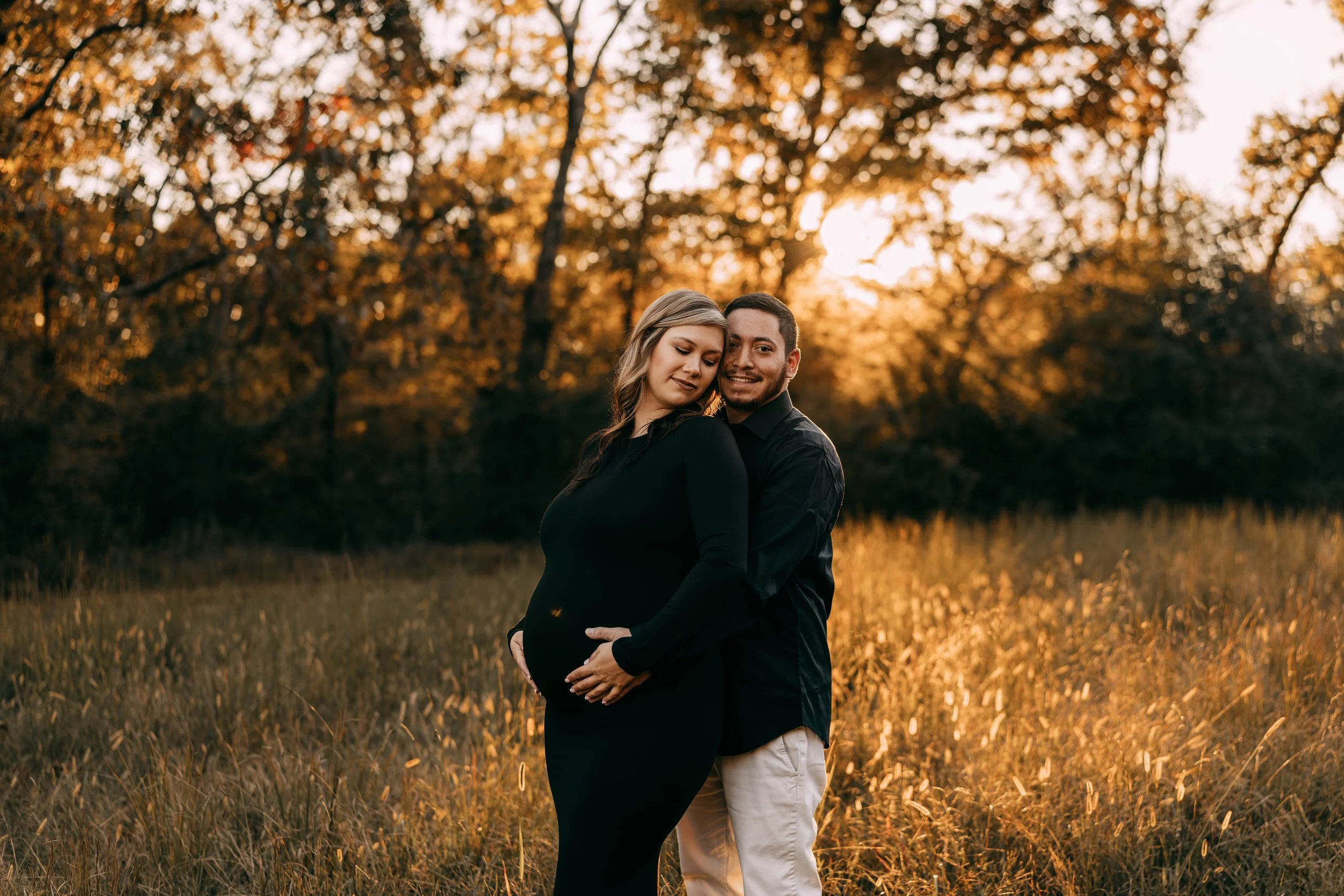 A pregnant woman in a black dress standing in a field with a man in a black shirt and beige pants during sunset, surrounded by trees with autumn leaves.