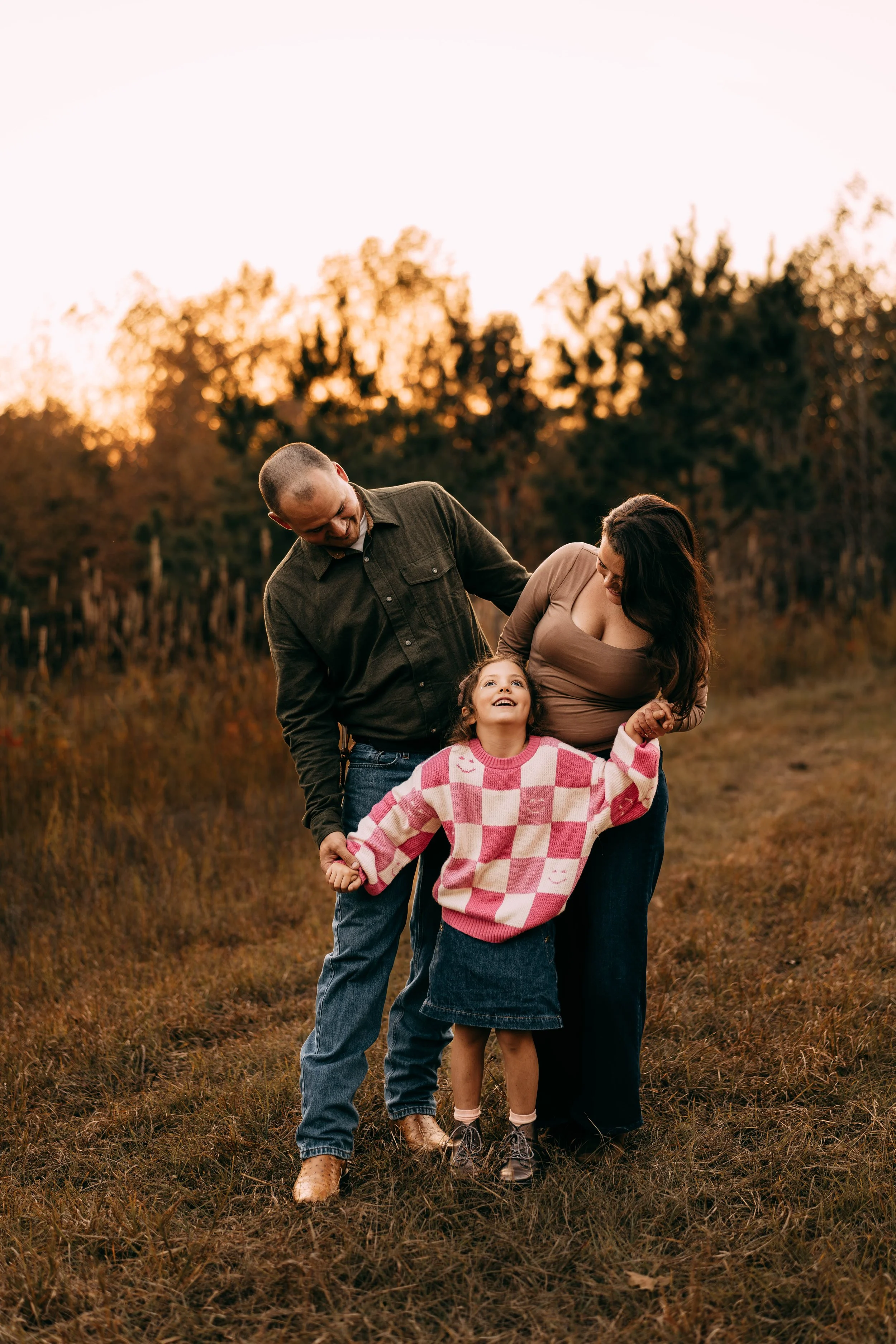 A family of three, a man, woman, and young girl, walking and laughing outdoors at sunset.