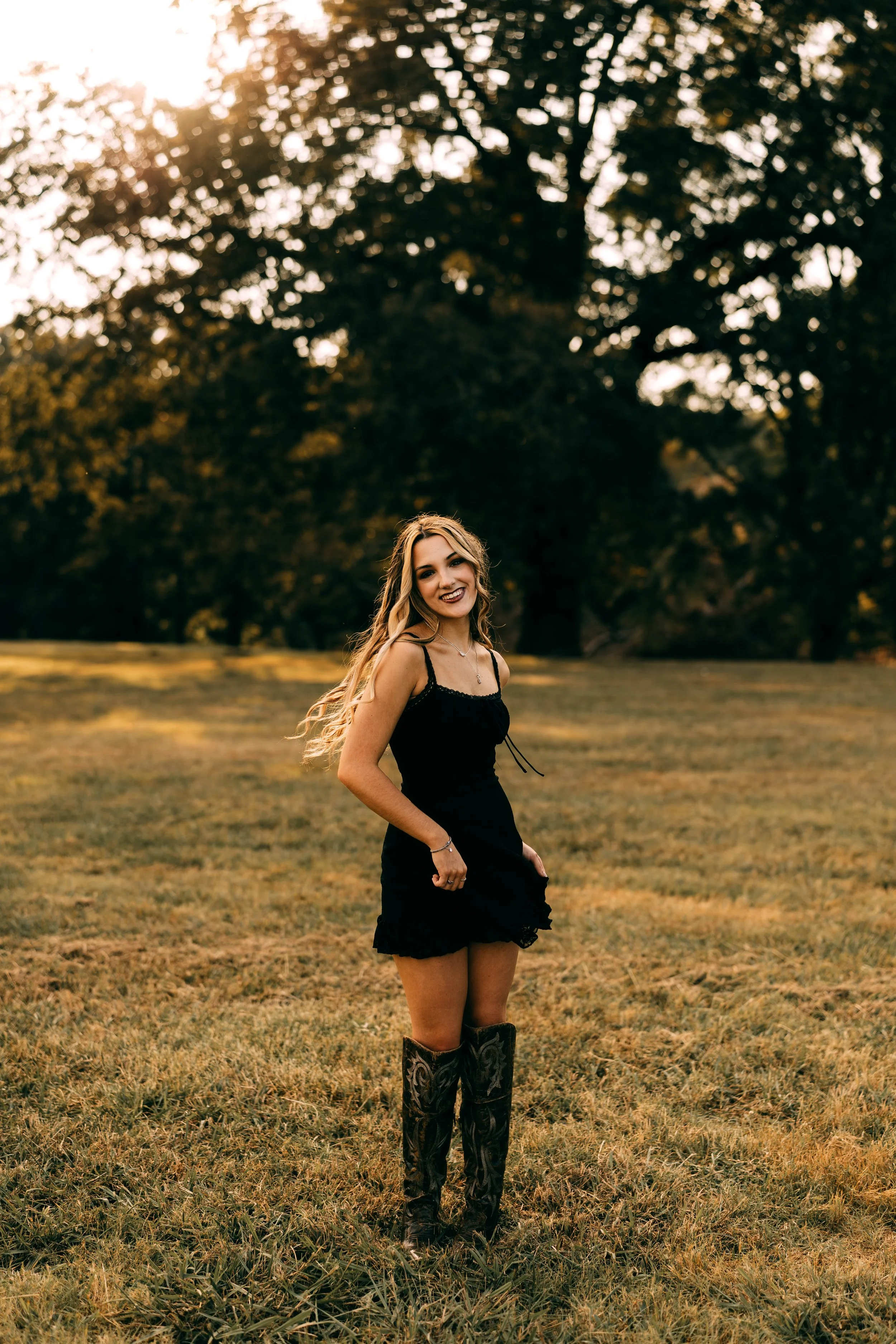 A young woman with long hair, wearing a black dress and cowboy boots, standing in an open field with trees in the background during sunset.