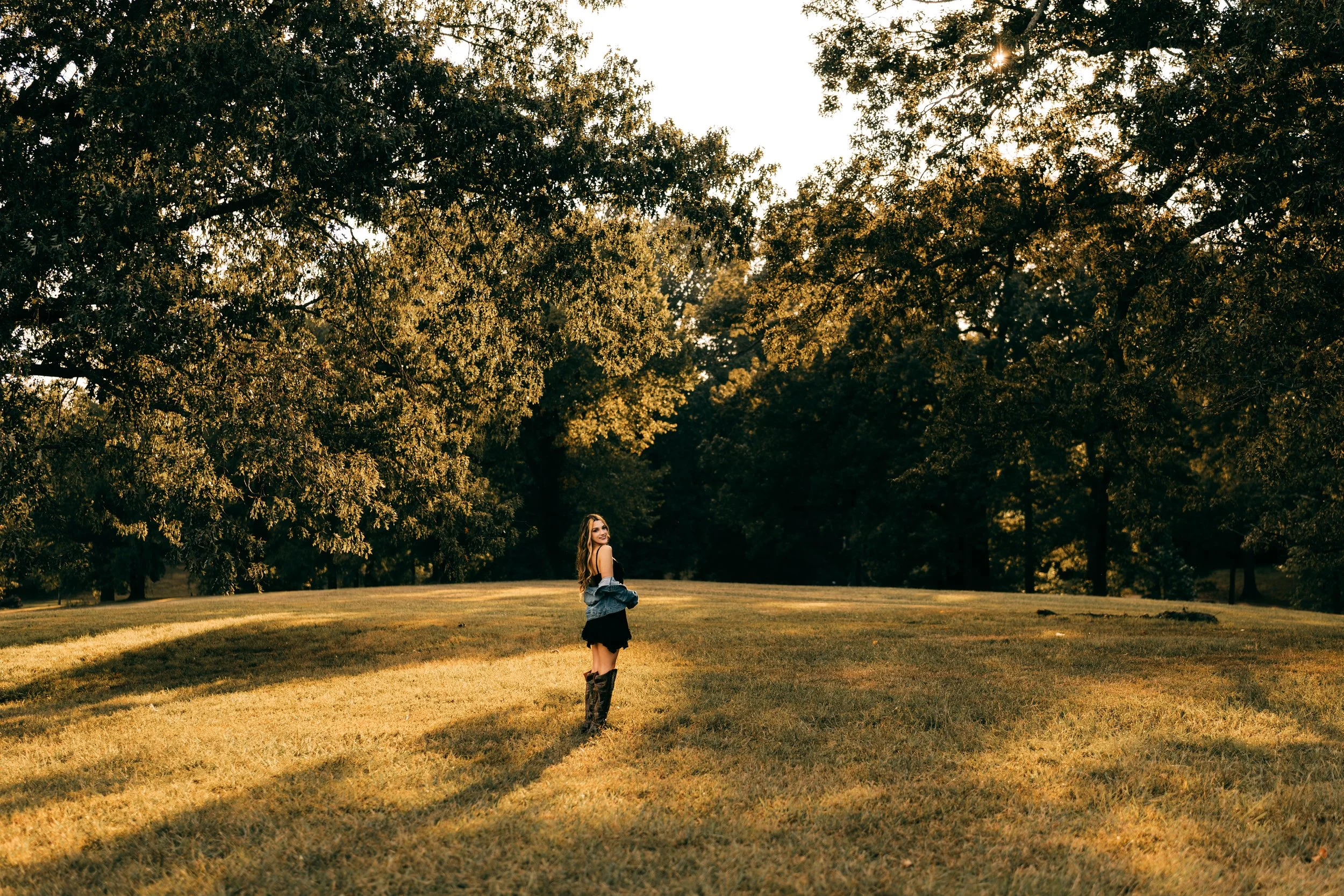 A woman standing alone on a grassy field during sunset, surrounded by large trees with golden leaves.