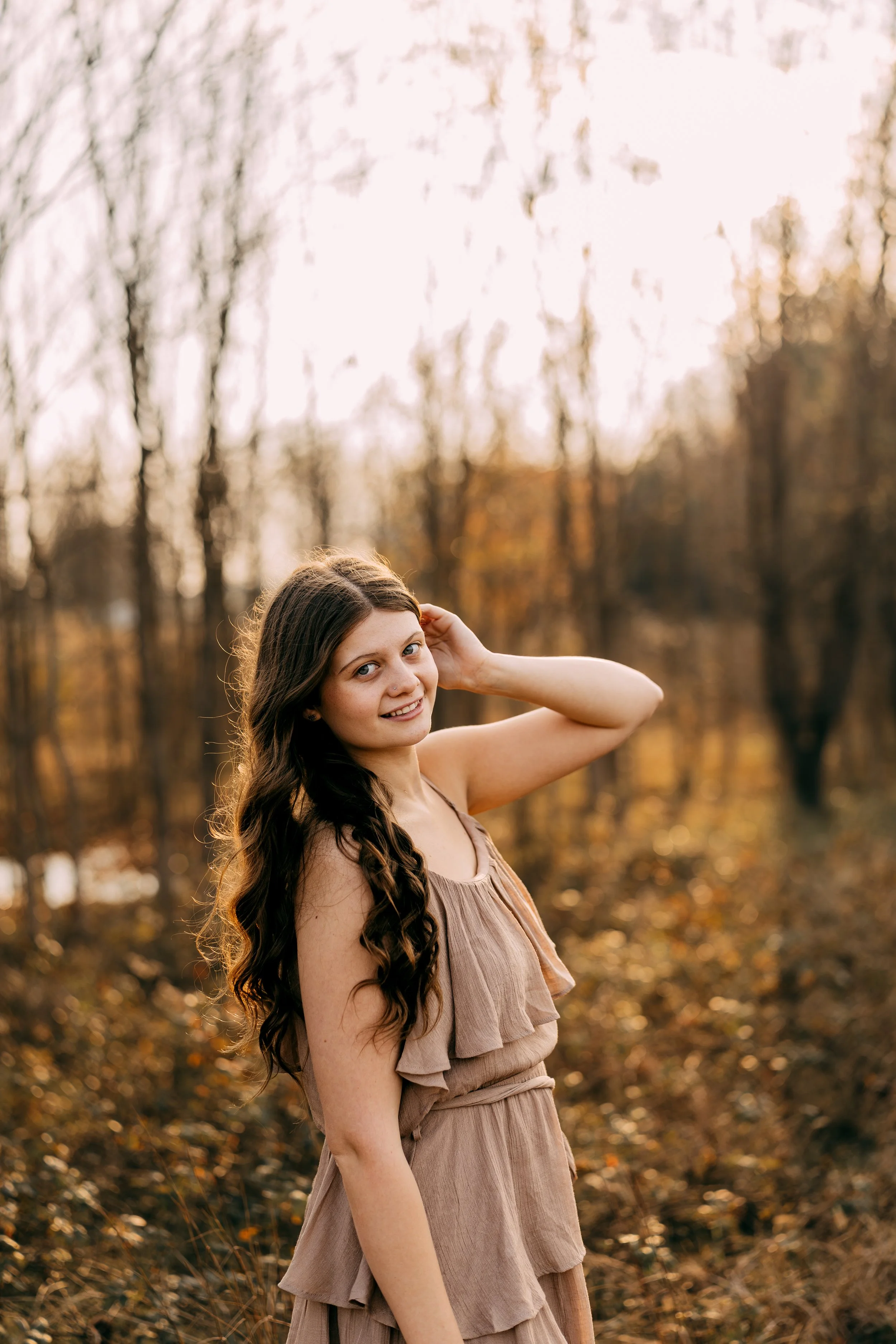 A young woman with long, wavy brown hair wearing a beige, sleeveless dress standing outdoors in a wooded area during autumn. She is smiling slightly, with one hand behind her head, and the background features bare trees and fallen leaves.