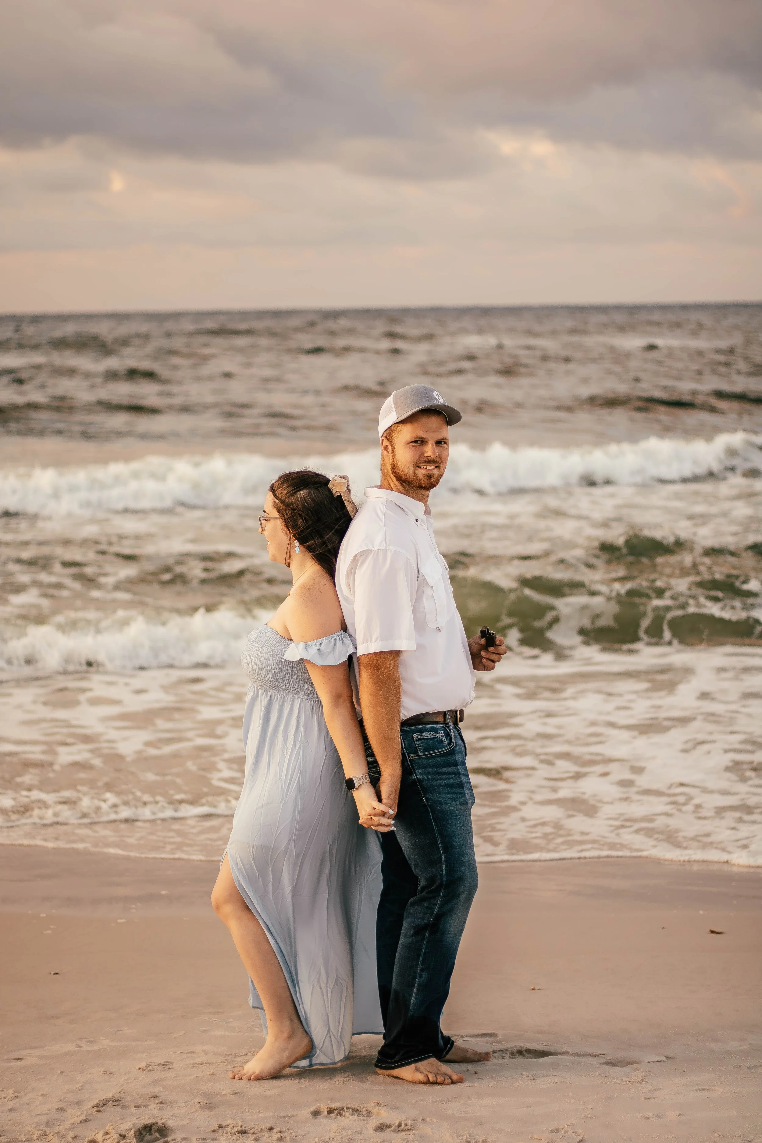 A couple holding hands on the beach, standing back-to-back near the shoreline with waves, during sunset.