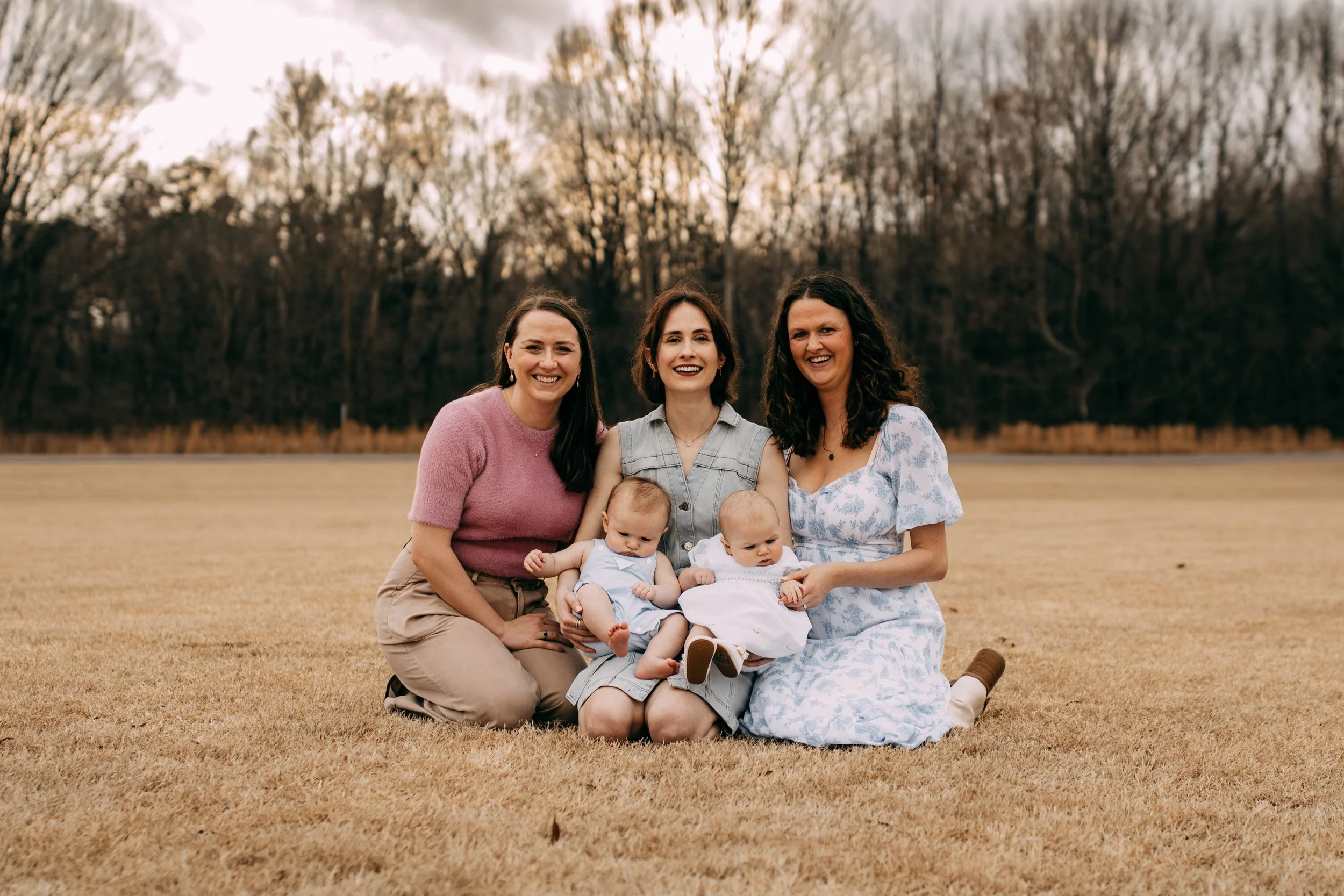 Three women sitting on a grassy field outdoors with two babies, all smiling. The background has trees with bare branches, suggesting late fall or early spring.