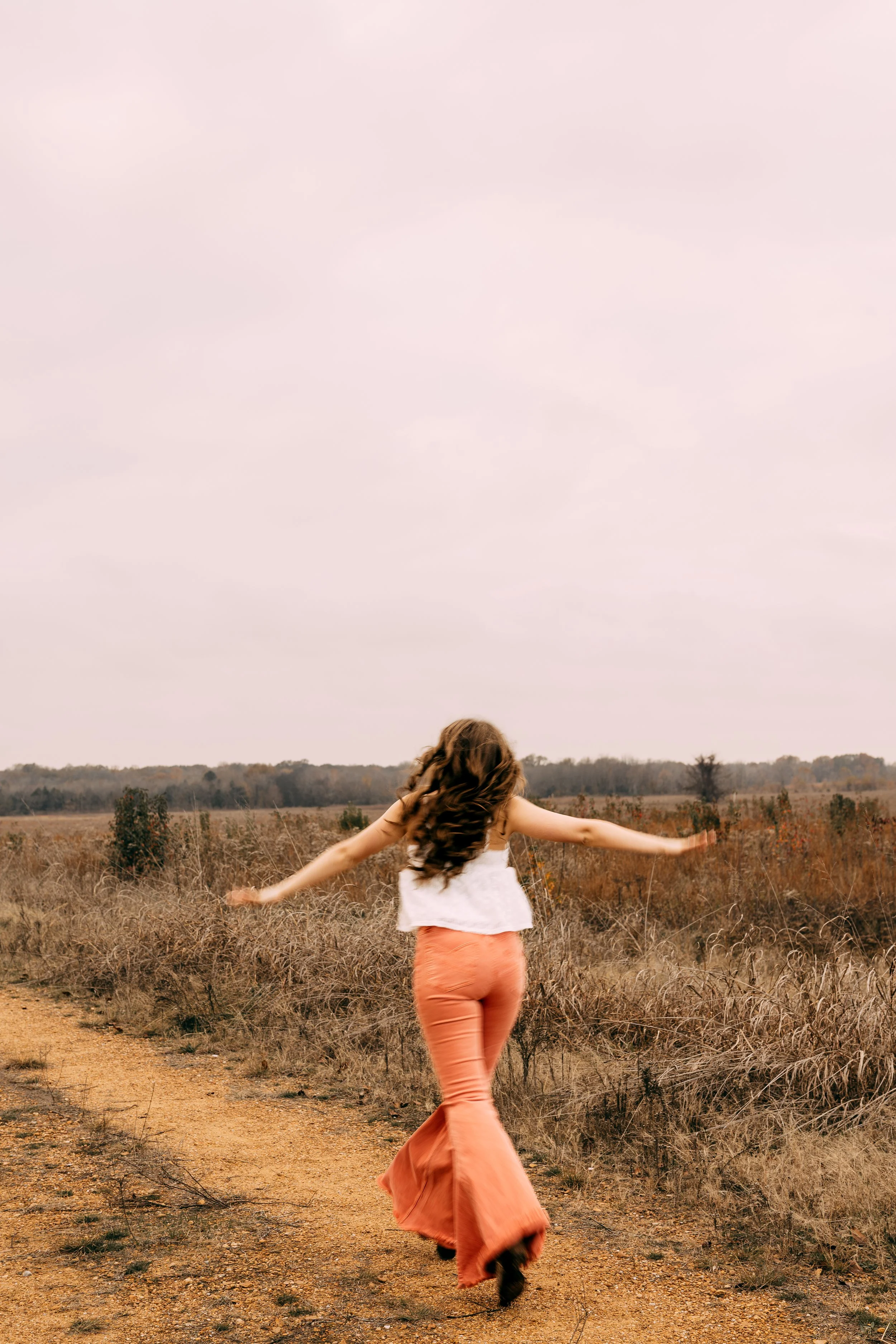A woman with long wavy hair wearing a white sleeveless top and long orange skirt walking along a dirt path in a field with dry grass, under a pale pink cloudy sky.