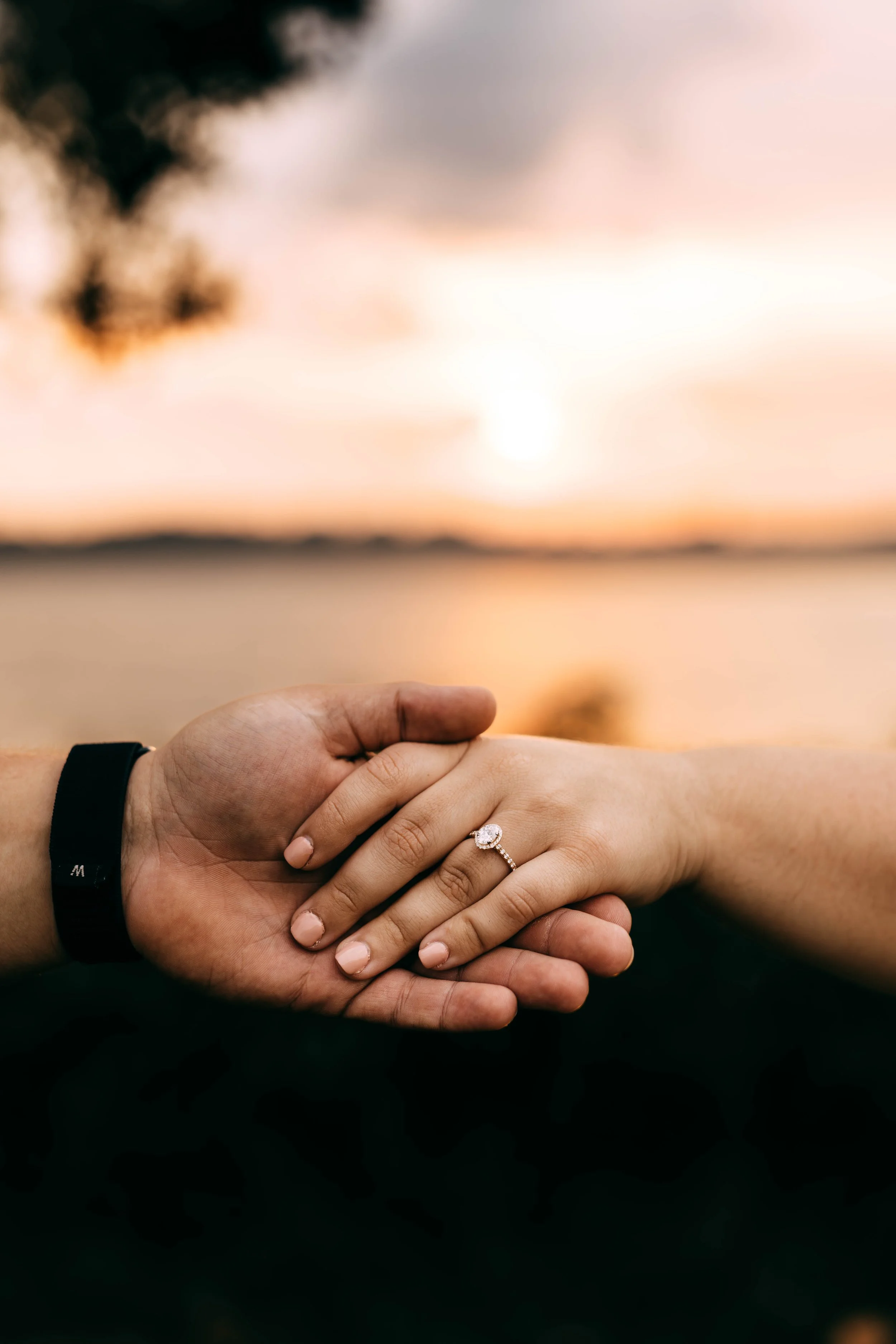 Close-up of a couple holding hands with an engagement ring on the woman's finger, against a sunset over a body of water.
