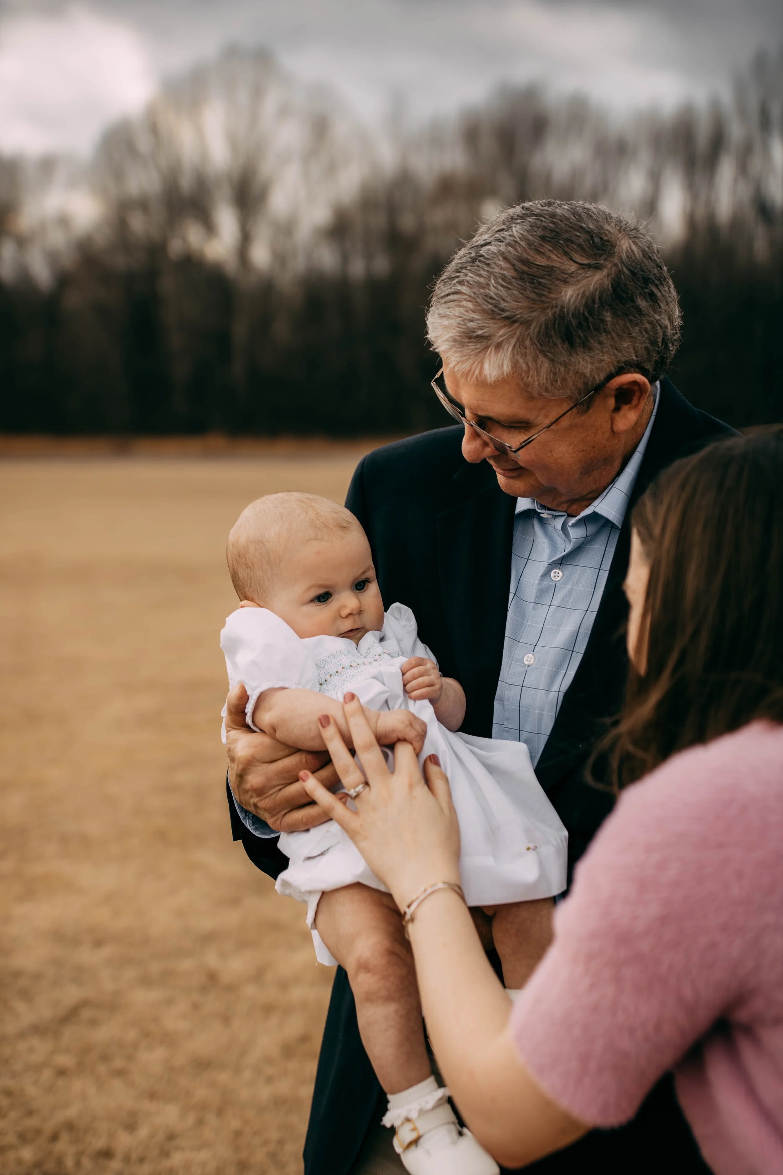 An older man holding a baby girl while a woman touches her hand outdoors with trees and a cloudy sky in the background.
